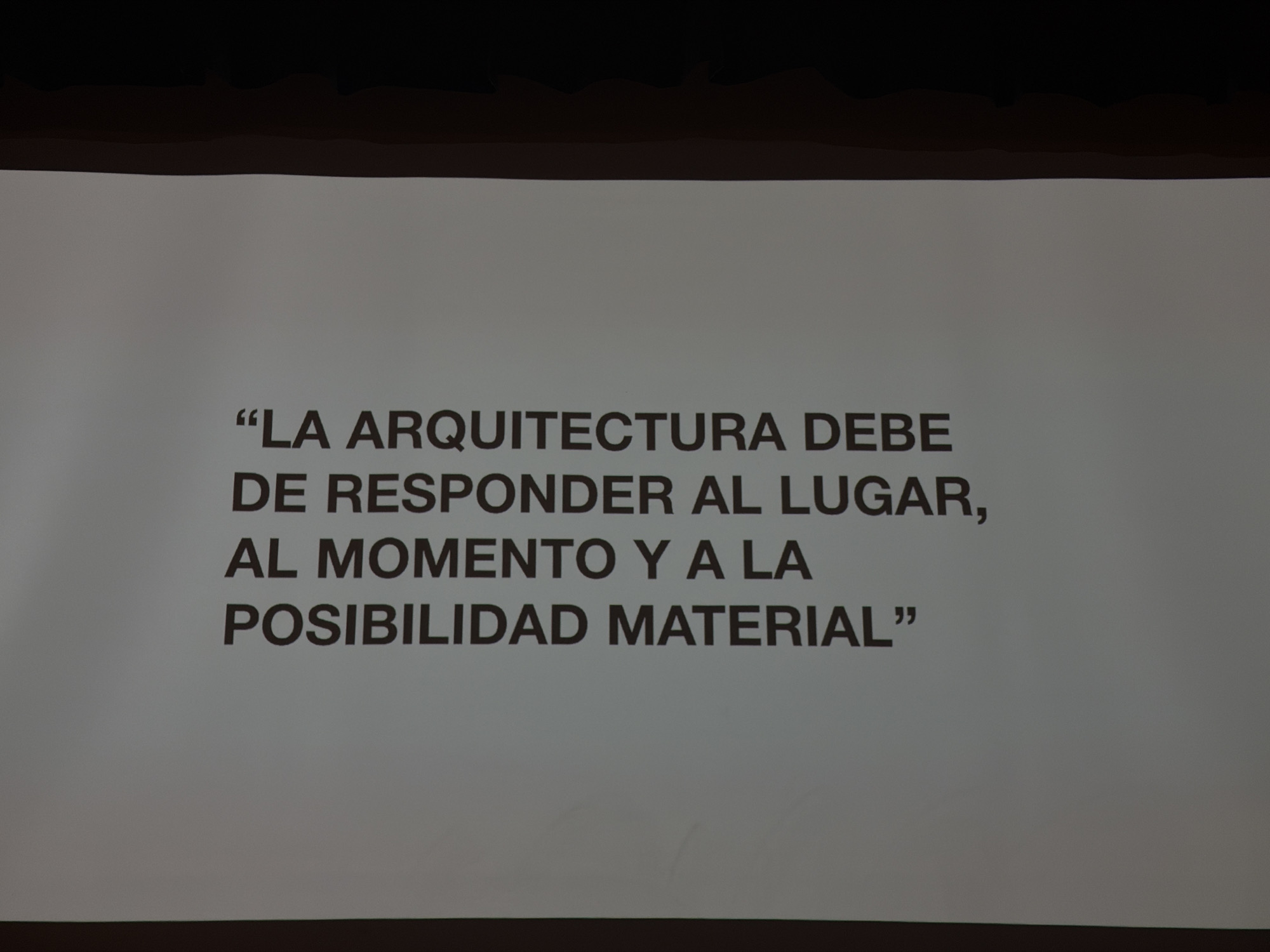The image shows a light grey wall with a dark grey top and bottom border. Printed on the wall in dark grey font are the following words, enclosed in quotation marks: "LA ARQUITECTURA DEBE DE RESPONDER AL LUGAR, AL MOMENTO Y A LA POSIBILIDAD MATERIAL".