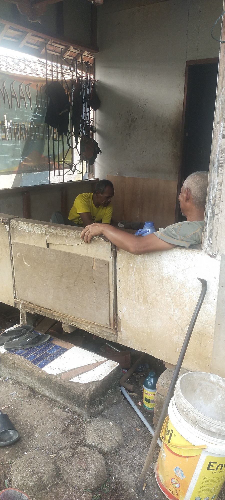 The image depicts an outdoor scene at a market stall in Pasirhuni, Indonesia. Two men are present. One man, wearing a yellow t-shirt, is seated and appears to be looking down. The other man, wearing a grey t-shirt, is leaning on a counter and looking towards the seated man. A blue bottle is visible on the counter. In the foreground, there are black flip-flops on a concrete step, and a large yellow paint bucket is positioned near a metal rod. The stall itself appears to be made of weathered wood and metal. Bags and other items hang from a grate behind the seated man, suggesting merchandise. The ground is uneven and appears to be dirt and gravel. The overall impression is one of a humble, working environment.