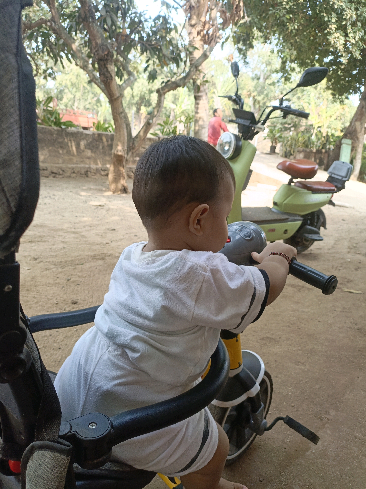 A child, viewed from behind, sits in a stroller and appears to be reaching for the handlebars of a parked electric scooter. The child is wearing a white outfit with black trim. The scooter is light green and has brown seats. In the background, trees and foliage suggest an outdoor setting. A person in a red shirt is visible in the distance. The ground is sandy.