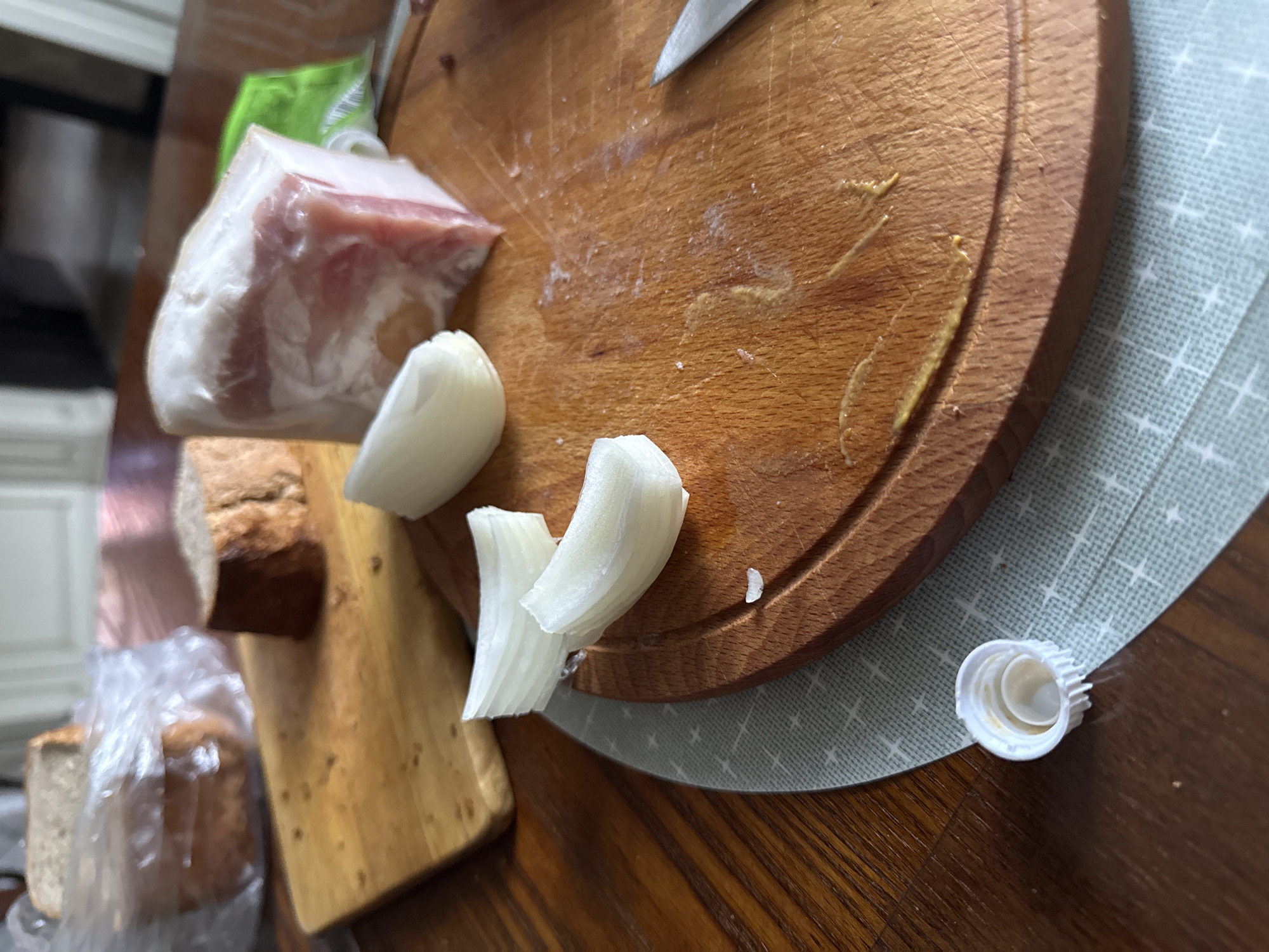 A medium shot captures a close-up of a rustic food preparation scene on a wooden table, likely in a kitchen or dining area in Krapivka, Russia. The primary focus is a well-worn round wooden cutting board, featuring several wedges of white onion and the tip of a knife blade. To the left of this board, a rectangular wooden board is partially visible, holding a block of what appears to be pork fat or lard (salo) with a pinkish layer of meat, still partially encased in clear plastic wrap. Further to the left, several slices of dark brown bread are visible, with some appearing to be in a clear plastic bag.
A light green placemat with a subtle white grid pattern is partially visible under the round cutting board and extending onto the wooden table, which displays a distinct wood grain. In the lower right corner of the frame, a small, white ridged plastic bottle cap lies on the placemat. The arrangement of ingredients suggests the preparation of a traditional snack or meal, possibly involving salo, onion, and bread, which are staples in Eastern European cuisine. The scene appears to be illuminated by ambient daytime light. There are no visible people or discernible text in the image.
