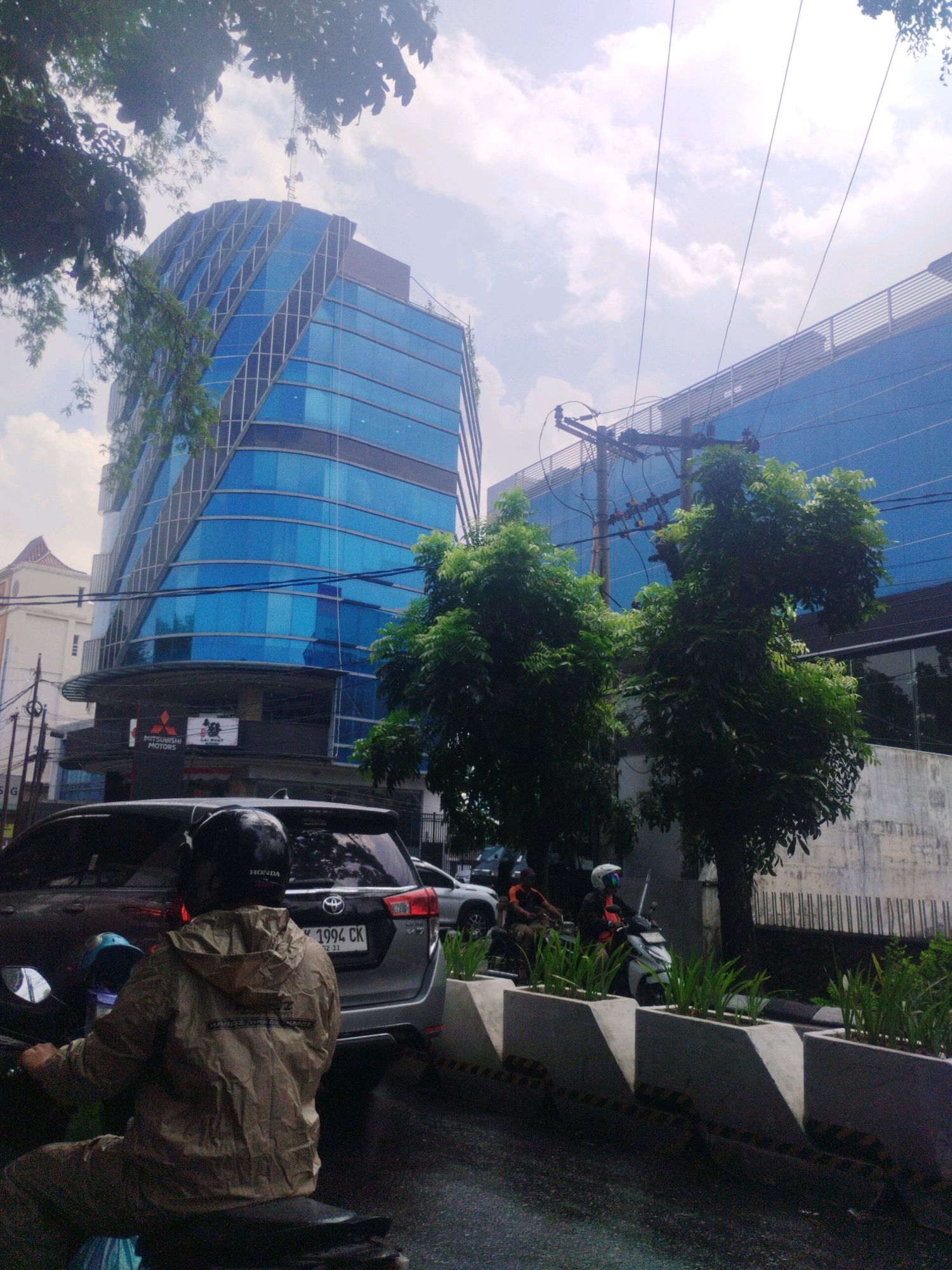The image captures a street scene in the City of Medan, Indonesia, dominated by modern architecture and urban elements. Two prominent blue glass buildings with distinctive angular designs stand tall, reflecting the cloudy sky. In the foreground, a person wearing a beige raincoat and a black helmet is riding a motorcycle, their back to the camera. To their right, a dark-colored car with a license plate reading "1994 CK" is visible.
A black and white power pole with multiple wires stretches across the frame, partially obscuring the buildings and sky. Lush green trees with dense foliage are scattered throughout the scene, adding a touch of nature to the urban landscape. On the right side of the image, a row of concrete planters filled with green plants lines the street. Another motorcycle with two occupants is seen further down the road, moving away from the viewer. The road appears wet, suggesting recent rain. The presence of the Mitsubishi Motors logo on a building facade indicates a commercial area.