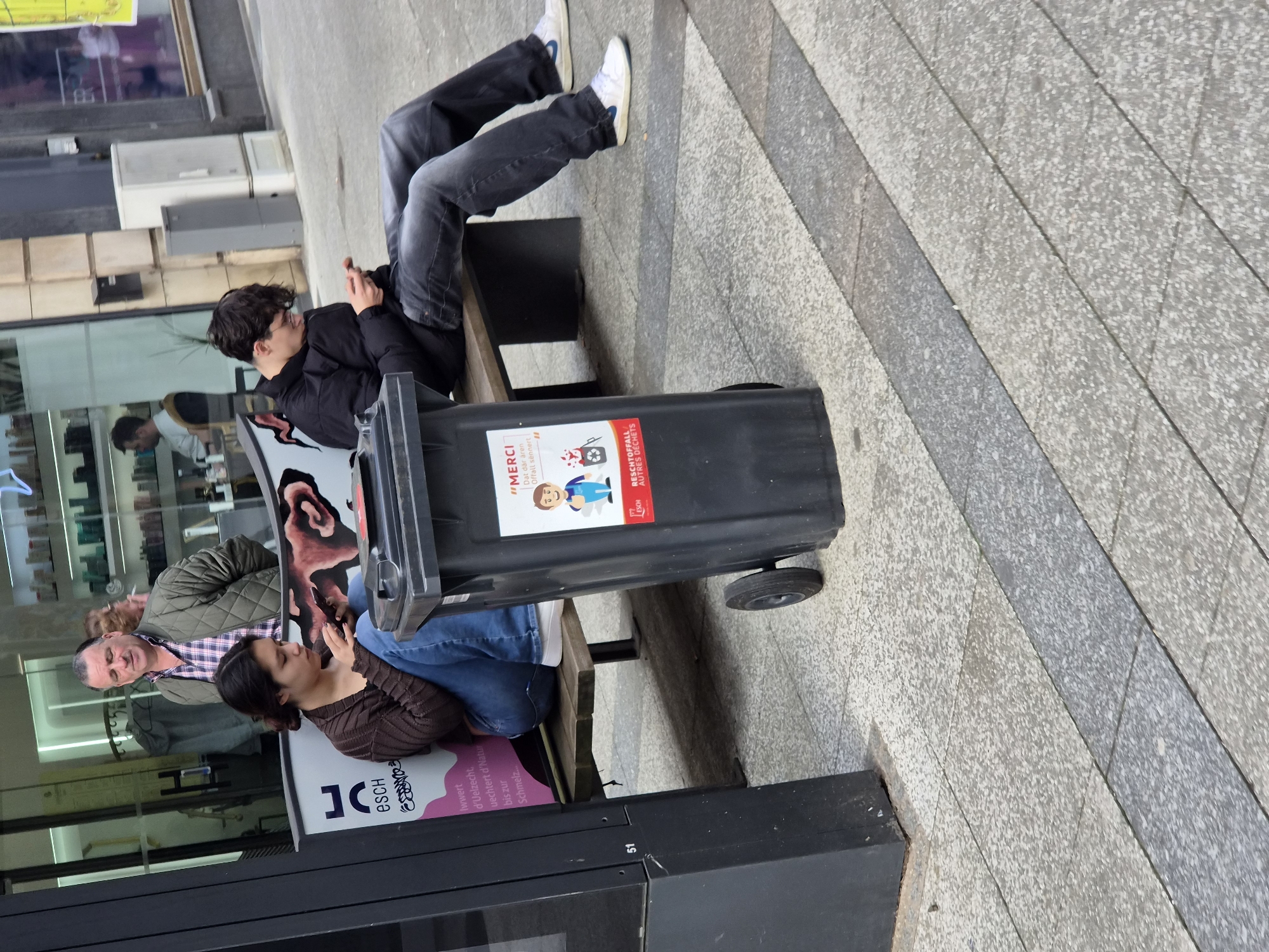 The image, taken in Esch-sur-Alzette, Luxembourg, depicts a daytime urban street scene. Three individuals are gathered around a wooden bench on a paved sidewalk next to building storefronts. The ground features a pattern of alternating light and dark gray textured tiles.
In the foreground, a young man with dark, curly hair and glasses, wearing a black jacket and dark jeans, sits on the bench with his legs extended, engrossed in a mobile phone. A large, black, wheeled trash bin separates him from a young woman also seated on the bench to his right. She has dark hair, wears a brown ribbed long-sleeve top and blue jeans, and is similarly focused on a phone in her hands. Behind her, an older man with lighter hair, dressed in a checkered shirt and a green quilted jacket, stands, looking towards the left. Reflections of other people and interior spaces are visible in the glass storefronts, suggesting a bustling environment.
The lighting is soft and diffused, indicating an overcast day with no harsh shadows. The overall atmosphere appears casual, with the younger individuals engaged with their devices.
Visible text includes information about waste sorting on the trash bin: "MERCI Dat där ären Offall sennert" (Thank you for sorting your waste) in Luxembourgish, and "ESCH RESCHTOFFALL AUTRES DÉCHETS" (Esch waste, other waste) in French/Luxembourgish, accompanied by an illustration. A pink sign, partially reflected in the storefront glass, shows Luxembourgish text: "esch Iwwert d'Uelzecht, vuechtert d'Natur bis zur Schmelz...", referencing the city Esch-sur-Alzette, the Alzette river, nature, and the historic "Schmelz" (steelworks). The number "51" is visible on a dark vertical panel at the bottom left, and some Japanese Katakana characters "ココ" are faintly visible in a reflection.