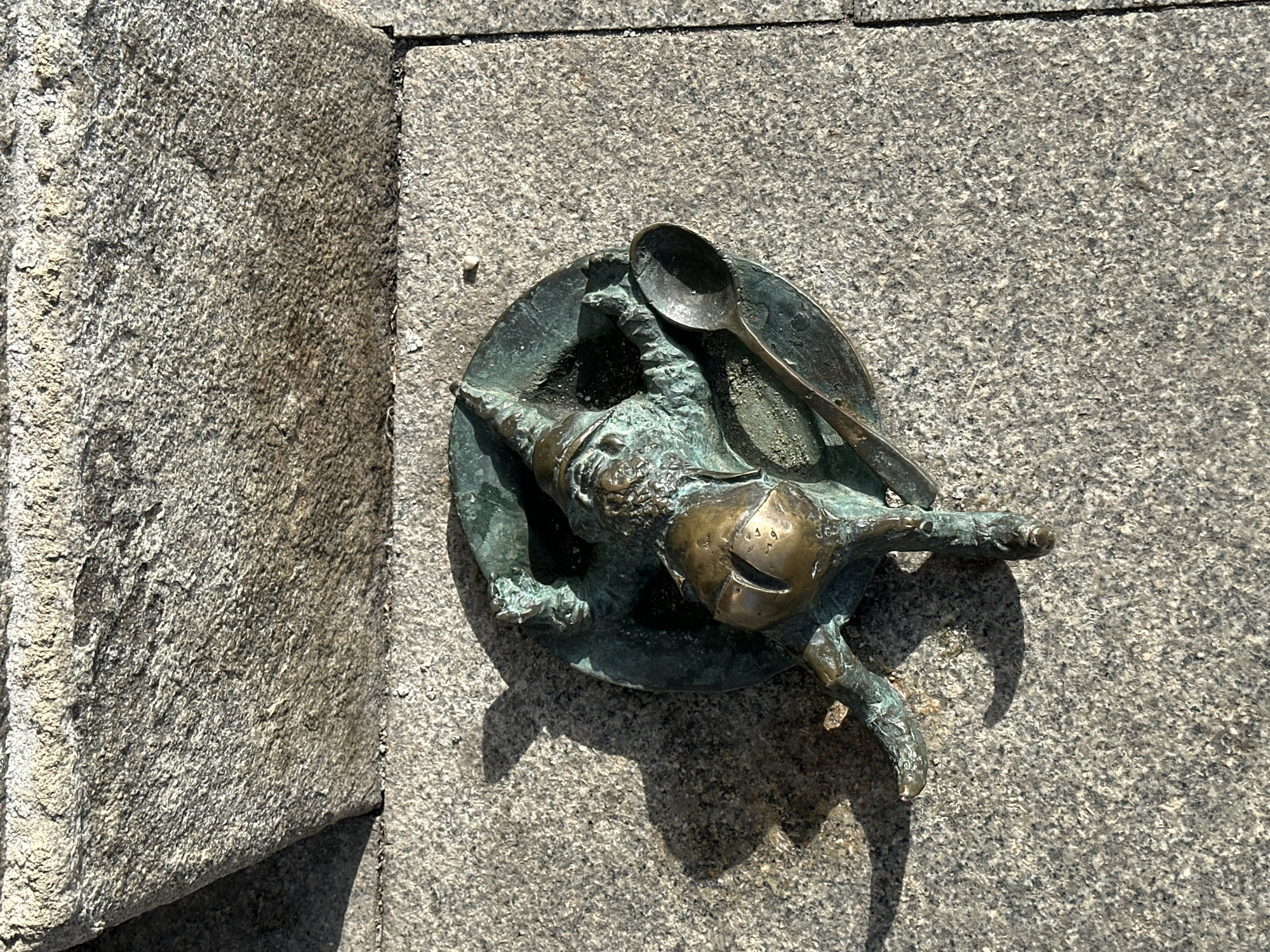 A high-angle medium shot depicts a bronze statue of a gnome on a plate with a spoon lying on a stone surface in Wrocław, Poland. The gnome statue is lying on its side on the plate. The stone surface is light gray with a fine, granular texture. To the left of the gnome is a corner of a larger stone structure. The statue and the stone surface both cast distinct shadows, suggesting a strong light source.