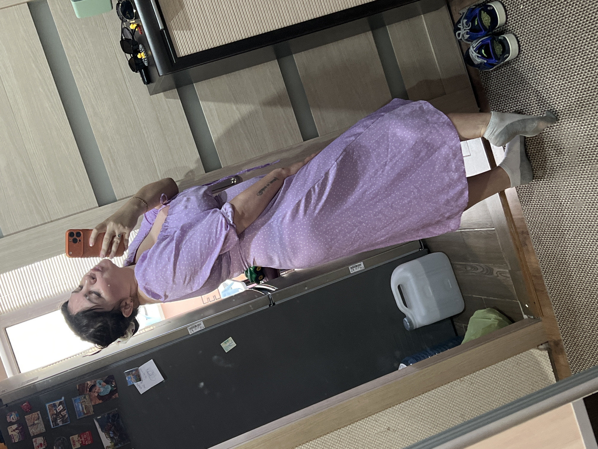 A young woman is taking a selfie in what appears to be a domestic environment. She is wearing a short-sleeved, purple polka-dot dress and light gray socks. Her hair is pulled back, and she is puckering her lips towards the camera. The background includes a refrigerator with magnets, a large white plastic jug, a green garbage bag, a wooden door, and a nightstand with sunglasses and other items. The floor is covered with a woven mat. The setting is likely an apartment or house interior. There is no visible indication of the specific city or location being Noviy, Russia.