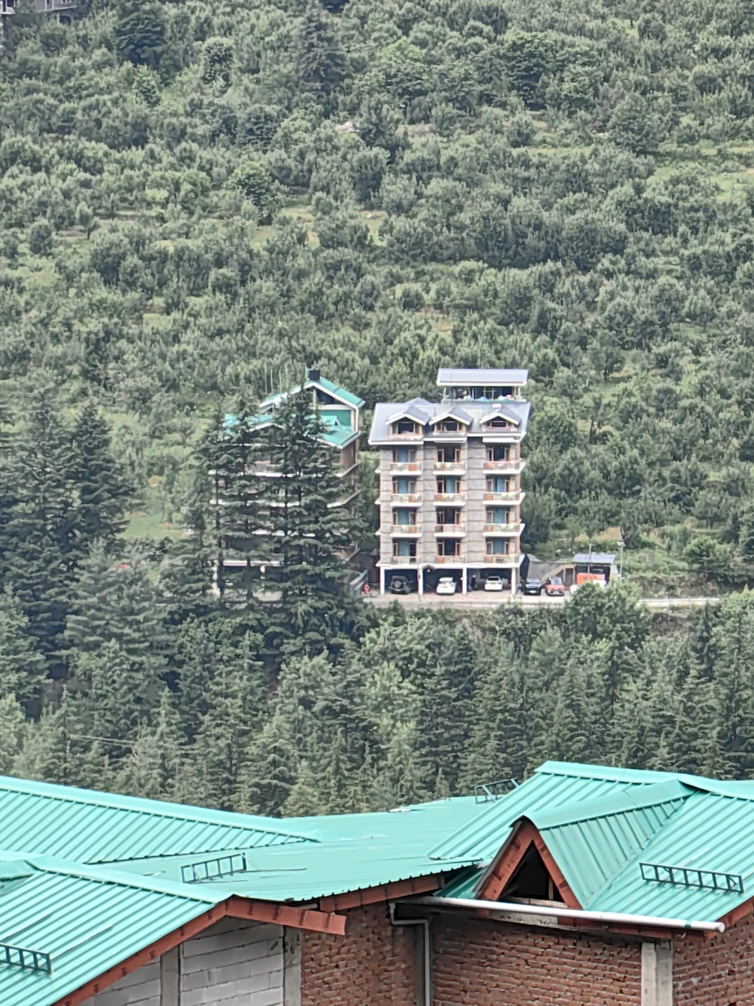 The image shows a multi-story hotel nestled on a densely forested hillside in Vashisht, India. The hotel is characterized by its gray concrete facade, multiple balconies, and a gray, multi-pitched roof with dormer windows. In front of the hotel, a road is visible with several parked cars, including a white sedan, a black SUV, and a red car. Adjacent to the hotel is another building with a green, sloped roof. The background is dominated by a vast expanse of lush green trees covering the undulating terrain, indicating a mountainous or hilly region. In the foreground, part of another building with a bright green, corrugated metal roof and brick lower walls is visible. The sky is not clearly depicted. The scene is devoid of any visible people or animals.