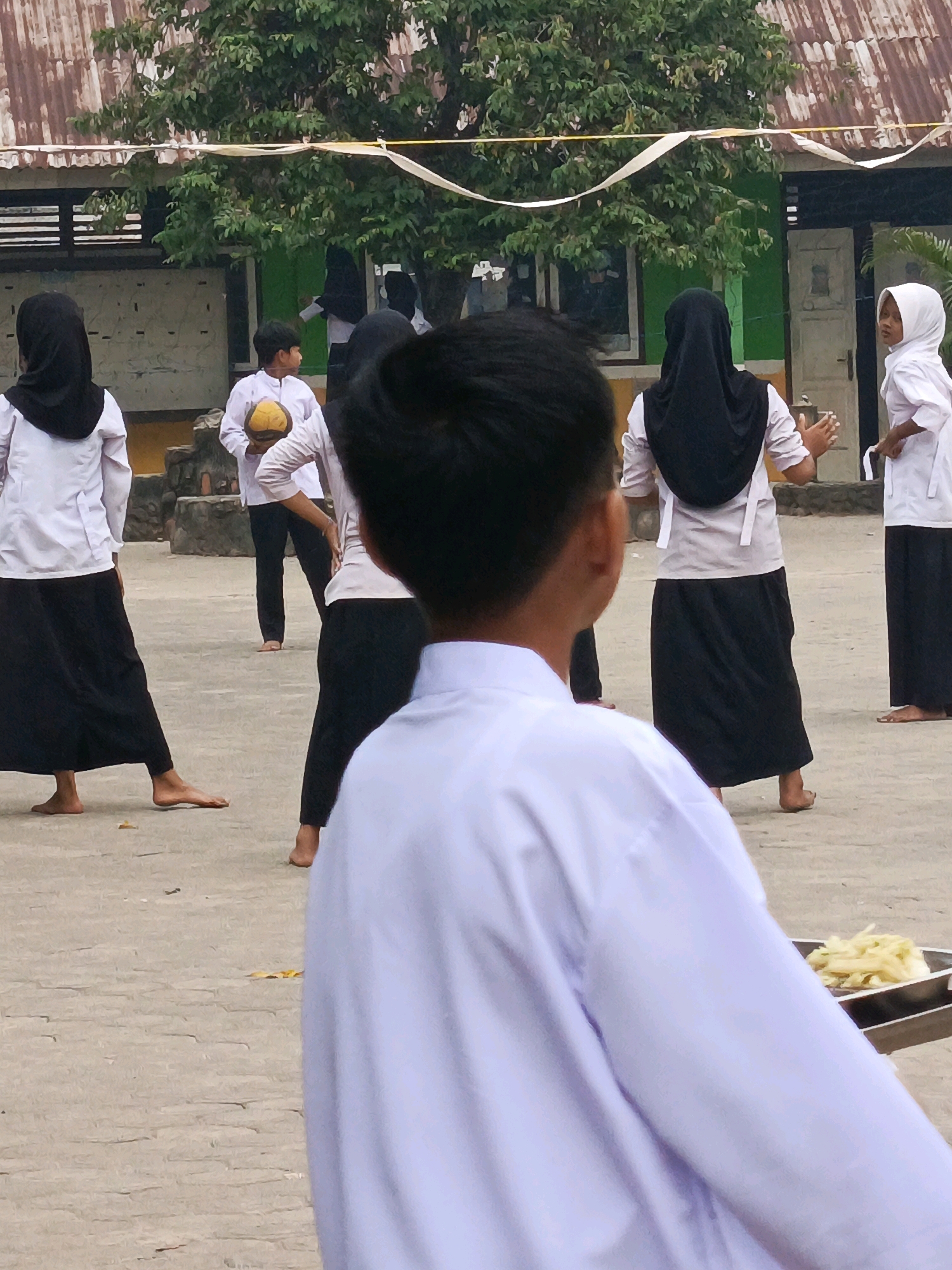 The image depicts an outdoor scene at an educational institution, possibly a school, located in Megang Sakti II, Indonesia. The foreground is dominated by the back of a person wearing a white long-sleeved shirt and possibly a white headscarf or veil, with their head turned away from the camera, looking towards a group of individuals in the mid-ground.
In the mid-ground, several young people, likely students, are engaged in an activity. Some are wearing white long-sleeved shirts and black long skirts or pants, along with headscarves or hijabs in black or white. One individual in the center is holding a yellow, round object, possibly a ball or a fruit. Another person to their right is wearing a white headscarf and a white top, with their hands extended forward. To the far right, another individual in a white headscarf and white shirt is visible. The ground is paved with a light-colored, textured material.
In the background, a building with a weathered, corrugated metal roof and green walls is visible. Trees with dense foliage are present, and some white streamers or ribbons are strung across the scene, possibly for decoration or an event. The overall atmosphere suggests a communal gathering or an outdoor lesson. The presence of food items in a metal tray in the lower right corner hints at a possible break or refreshment period.