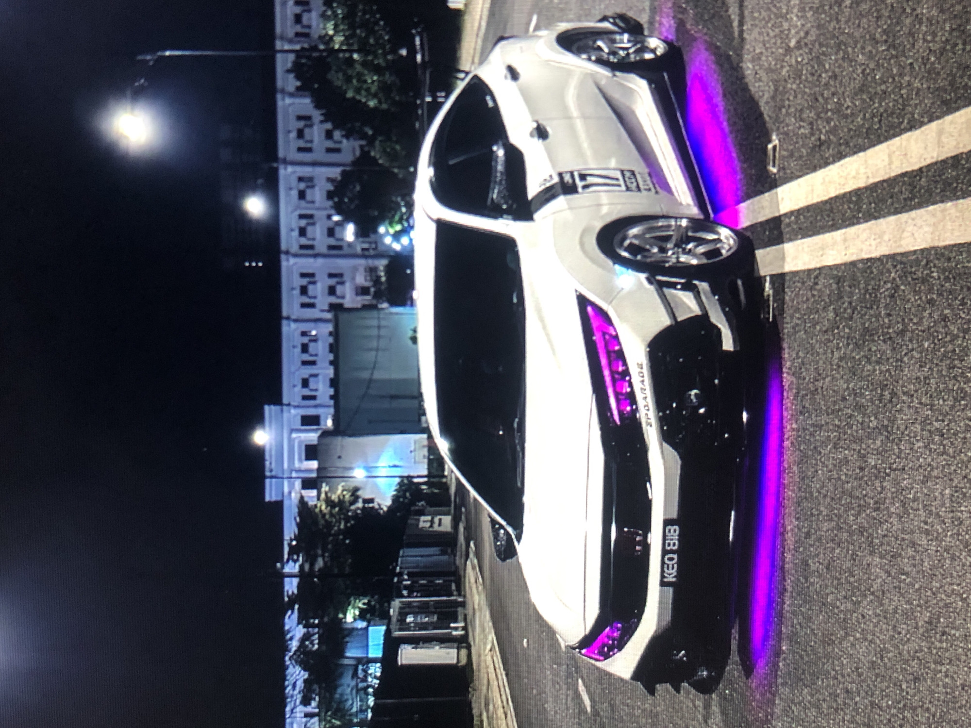 The image depicts a white car with purple neon lights parked on an asphalt road. The license plate reads "KEQ 818." The car has racing decals, including the number "17." A building with multiple windows is visible in the background, illuminated by street lights. This image was taken in Lhokseumawe, Indonesia, at night.