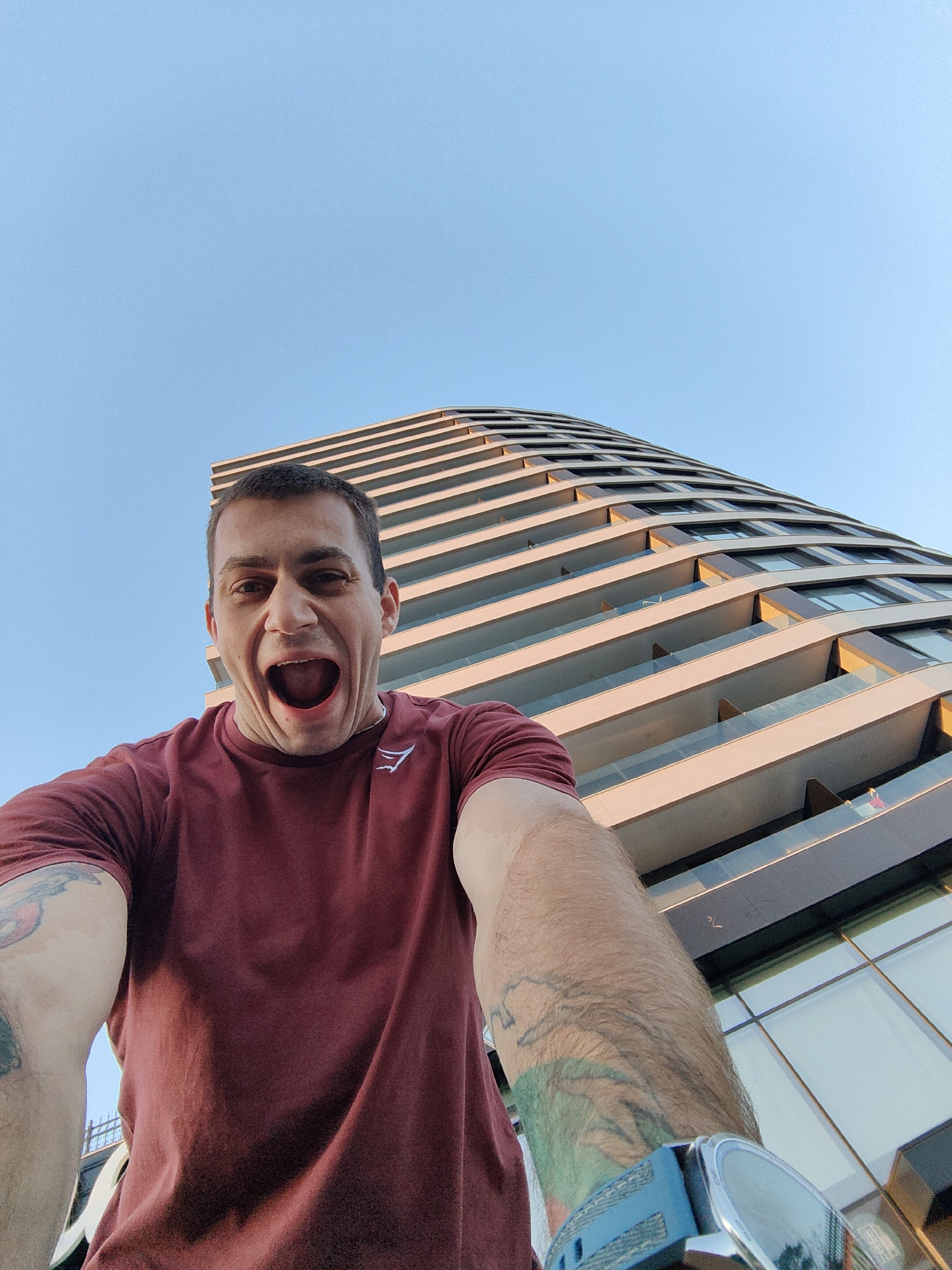 A low-angle, wide shot captures a man with short, dark hair and an open mouth, looking up at the camera against a clear blue sky. He is wearing a maroon t-shirt with a white logo on the left chest. A portion of a modern, curved building with many balconies and windows is visible in the background. The building appears to be an apartment or hotel complex. The scene is likely set in Varna, Bulgaria, given the context of the building's architecture, which is characteristic of contemporary developments in the city. The lighting suggests it is daytime, possibly late afternoon due to the warm glow on the building's facade. The man's expression is one of surprise or excitement.