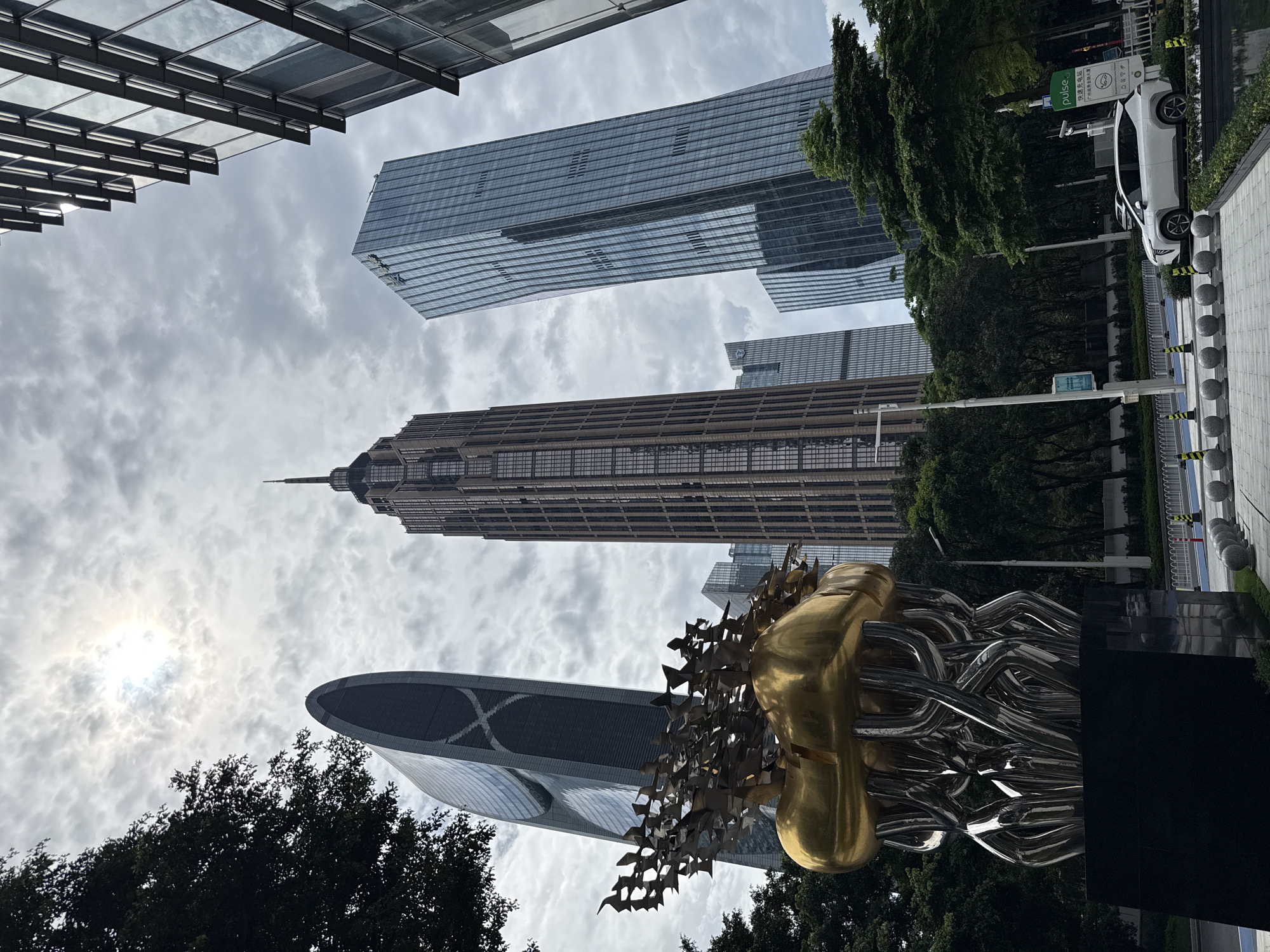 In Tianhe District, China, a public sculpture composed of abstract metallic forms supports a golden, bulbous element from which numerous bird-like figures are emerging. The sculpture is situated in front of a row of modern skyscrapers with glass and metal facades, under a cloudy sky. The tallest building is a brown skyscraper with a pointed spire. To its left is a unique, curved, dark-colored skyscraper with an 'X' pattern on its side. To the right of the brown skyscraper is a tall, slender blue glass skyscraper. In the foreground, a white SUV is parked near a charging station with a green sign that reads "pulse" and Chinese characters. The street is lined with trees, bollards, and decorative spheres.