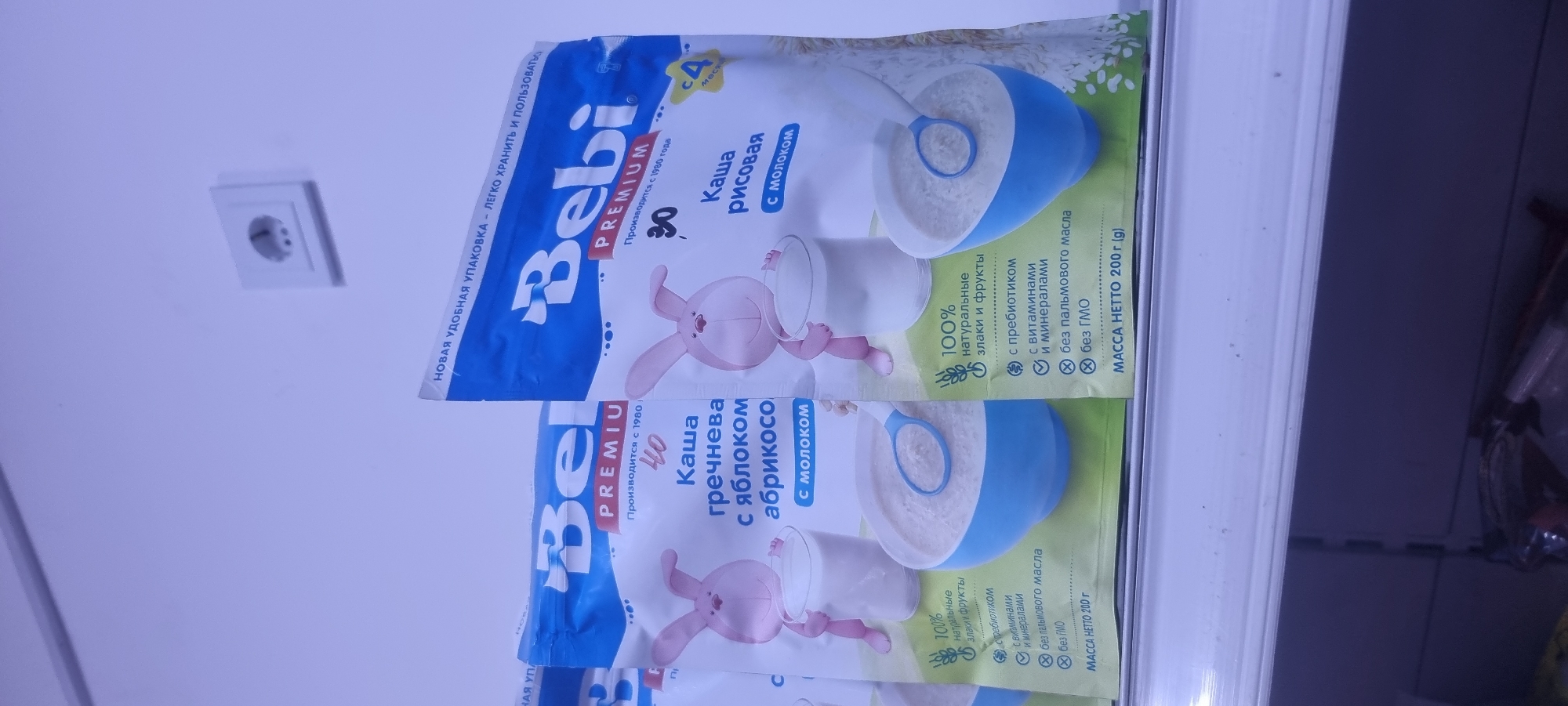 The image shows several packages of "Bebi" brand baby food, specifically powdered porridge. Two packages are prominently displayed on a white shelf. The package on the left is for "Buckwheat porridge with apple and apricot with milk," and the package on the right is for "Rice porridge with milk." Both packages feature illustrations of a pink rabbit holding a glass of milk, and a bowl of porridge with a spoon. Text on the packages indicates they are made with natural grains and fruits, contain prebiotics, vitamins, and minerals, and are free from palm oil and GMOs. The net weight of each package is 200g. The background is a light blue wall with a white electrical outlet. This image was taken in a retail environment, likely a grocery store or supermarket, in the city of Ismoili Somoni, Tajikistan. The overall scene is well-lit and the products are clearly visible, suggesting a staged photograph for advertising or inventory purposes.