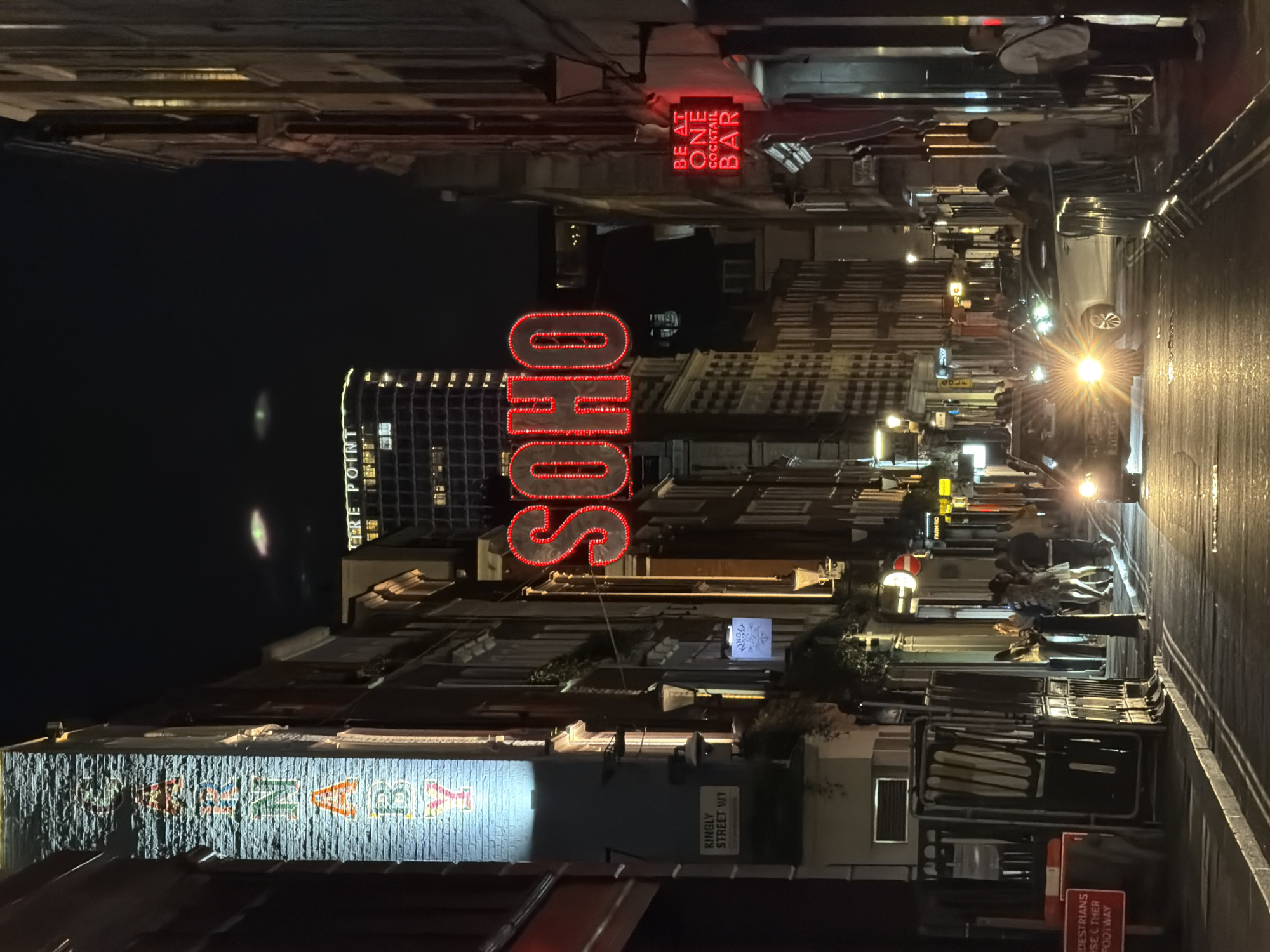 The image captures a street scene in Soho, London, at night. Visible are several illuminated signs, including the word "SOHO" in large red letters, "BE AT ONE COCKTAIL BAR," and "KINGLY STREET W1." A building is labeled "CTRE POINT." On the street are pedestrians, a car with illuminated headlights, and street furniture. A sign advises pedestrians to use the other footway. The environment appears to be urban, with closely packed buildings lining the street.