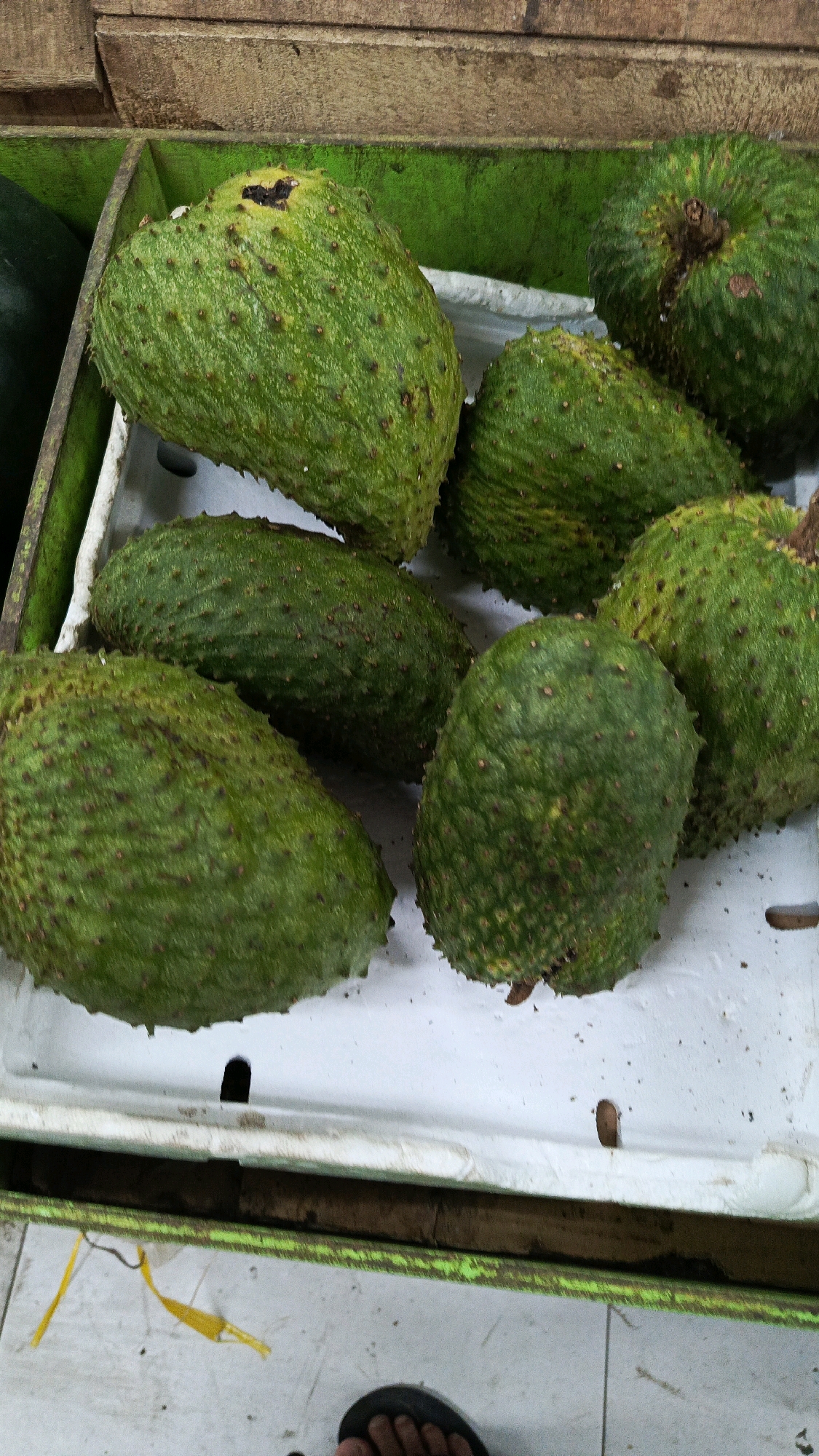 A close-up, overhead shot shows a collection of soursop fruits arranged in a green bin with a white liner. The bin is placed on a tiled surface in what appears to be an outdoor market or stall. The fruits are predominantly green with bumpy skin and some have brown spots. One fruit in the top left corner has a dark, damaged area. Wooden planks are visible at the top of the frame, suggesting the stall structure. A person's foot wearing a dark sandal is visible at the bottom right of the image, on the tiled floor. Yellow plastic ties are also present on the floor. The overall impression is a display of fresh produce.