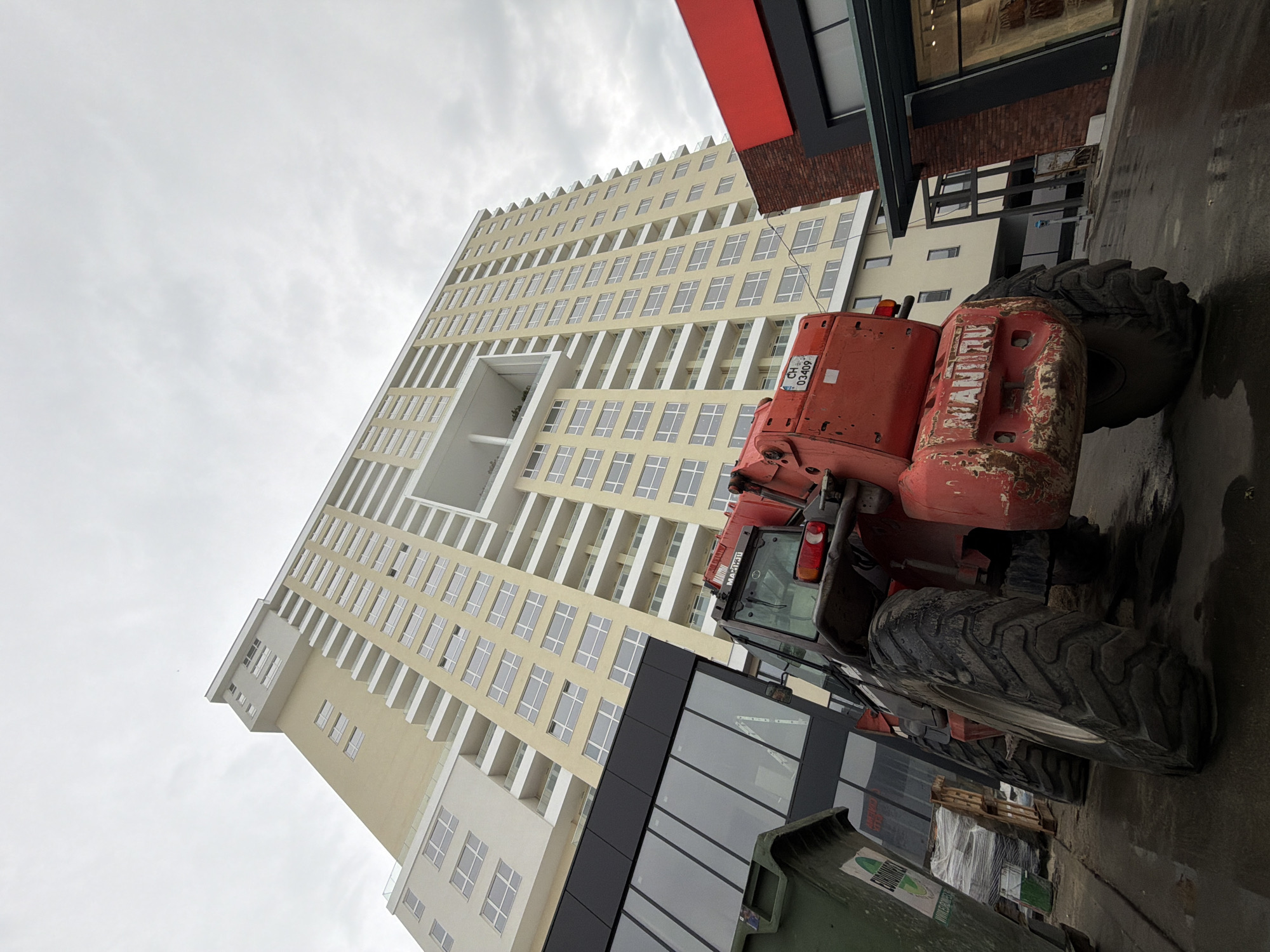 The image depicts a scene in Burgas, Bulgaria, featuring a tall, modern building under construction or renovation. The building is a light yellow color with numerous windows and white balconies, with a distinct architectural feature of a large open space near the top.
Positioned in front of the building is a red Manitou telescopic handler, a type of construction equipment used for lifting and moving materials.
In the foreground, there is a green dumpster and some wrapped pallets, suggesting an active construction or renovation site. The ground appears wet or slightly reflective, possibly from a recent rain. The sky is overcast, indicating a cloudy day.
The scene includes elements of both the urban landscape (the modern building) and the construction/industrial aspects (the Manitou and the site equipment).
