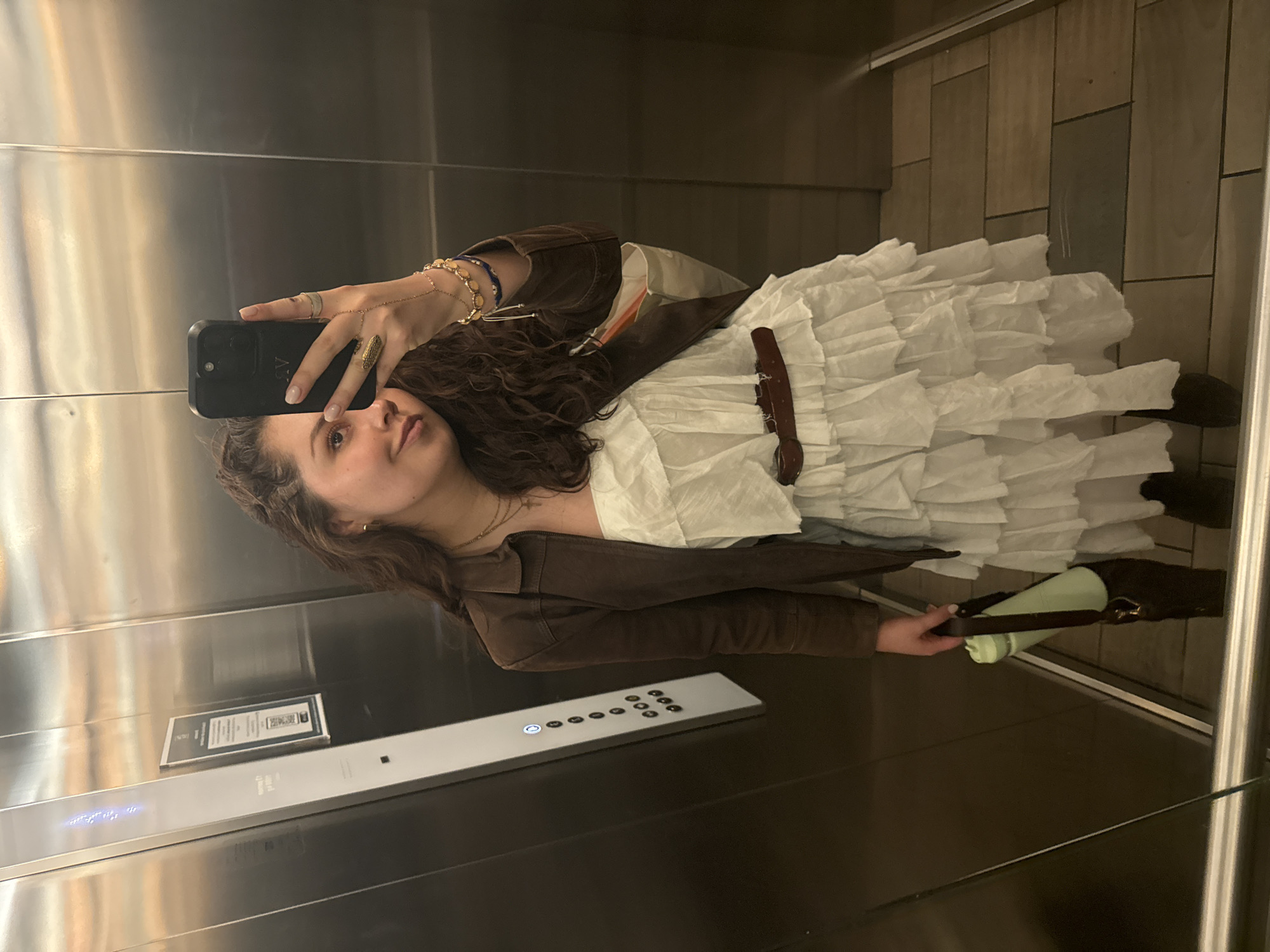 A young woman is taking a selfie in what appears to be an elevator. She is holding a smartphone in her left hand and looking at the screen. She is wearing a brown jacket over a white dress with multiple ruffled tiers, and several bracelets are visible on her left wrist. She also appears to be holding a green tumbler in her right hand.
The elevator interior is made of metal panels. An elevator control panel with illuminated buttons is visible. A sign with text and a QR code is mounted on the wall. The elevator floor is dark and there are tile-like panels in the upper right.
Based on the appearance and design of the elevator, it is likely that the photo was taken in Mexico City, Mexico. However, further details would be needed to confirm that.
