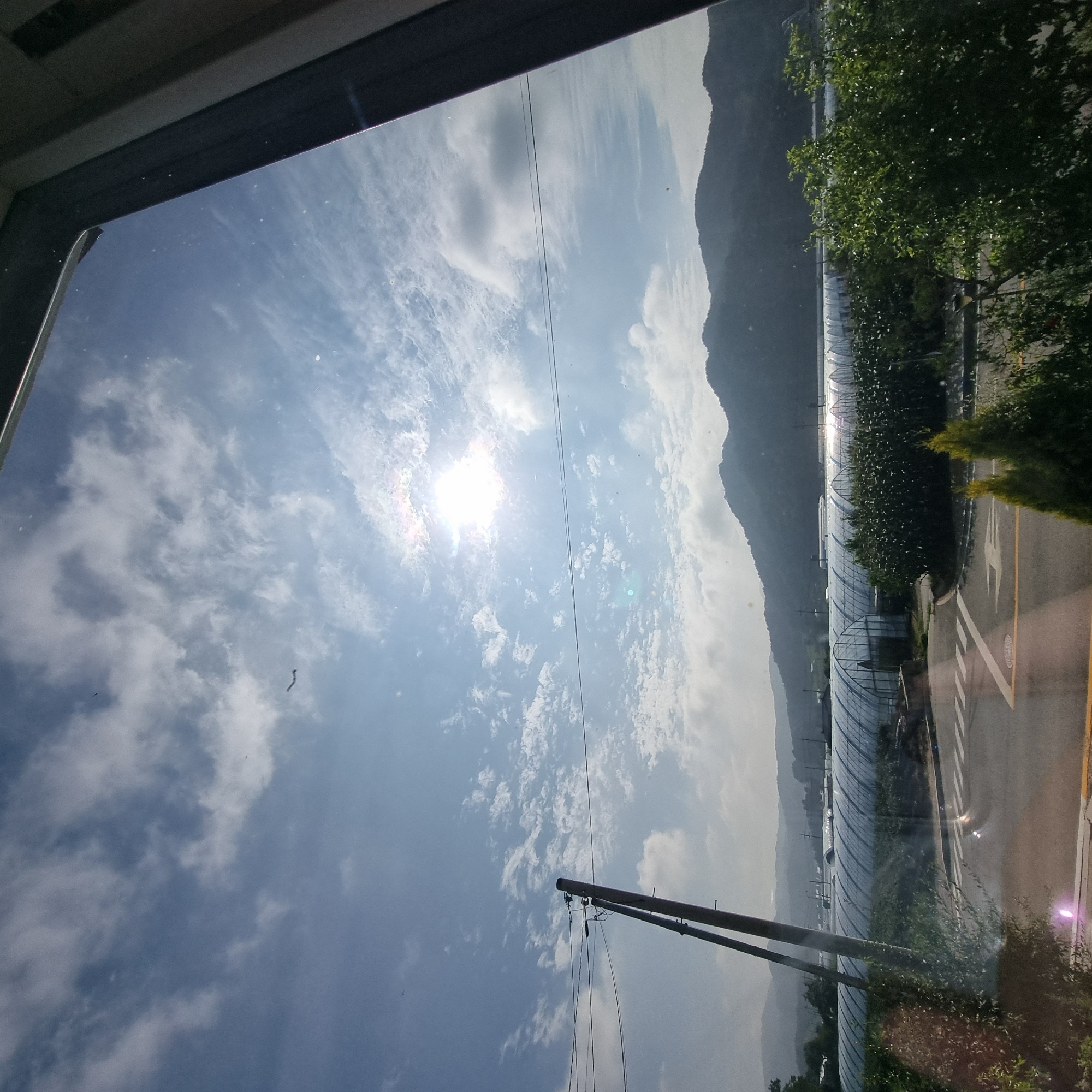 The image shows a wide shot of a rural landscape, likely in Dunnae-myeon, South Korea. In the foreground, a dark window frame occupies the right side and top edges of the image. A gray asphalt road with white lane markings and a pedestrian crossing is visible in the lower part of the scene. To the left of the road, a telephone pole with electrical wires is prominently featured. Behind the road and telephone pole, rows of translucent greenhouses stretch across the landscape. These greenhouses appear to be for agricultural purposes, possibly for growing crops. In the background, a range of dark, tree-covered mountains forms the horizon. The sky above is a bright blue, with scattered white clouds and the sun shining intensely, creating a lens flare effect in the upper middle of the image. The overall impression is of a sunny day in a cultivated rural area, viewed from indoors.