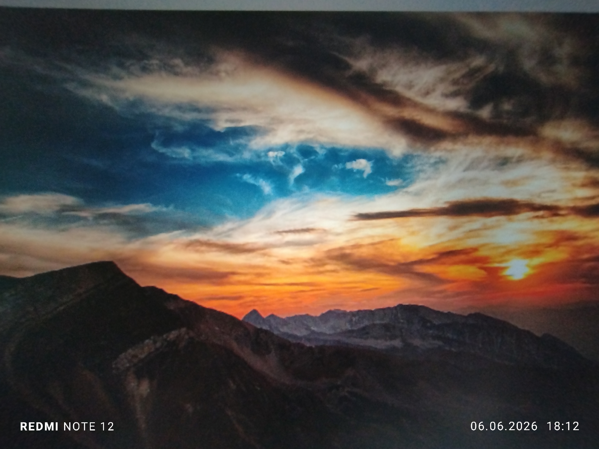 The image displays a panoramic mountainous landscape during either a sunset or sunrise. The sky in the upper left transitions from dark blue to lighter blue with scattered white and grey clouds. A distinct band of darker, elongated clouds extends across the upper third of the frame. Towards the upper right and middle right, the sky is illuminated by intense orange, red, and yellow light, with a visible, bright orange sun disk positioned low above the distant horizon.
Multiple layers of dark, silhouetted mountains dominate the lower half of the scene. The nearest mountain range on the left features rugged peaks and shadowed slopes. Subsequent mountain ranges recede into the distance, appearing progressively lighter and hazier. There are no identifiable people, animals, or man-made objects within the natural landscape.
Two text overlays are present: "REDMI NOTE 12" is located in the bottom left corner, and "06.06.2026 18:12" is displayed in the bottom right corner. The depicted mountainous environment is geographically inconsistent with the city of Burgas, Bulgaria, which is a coastal city not characterized by such prominent mountain ranges.