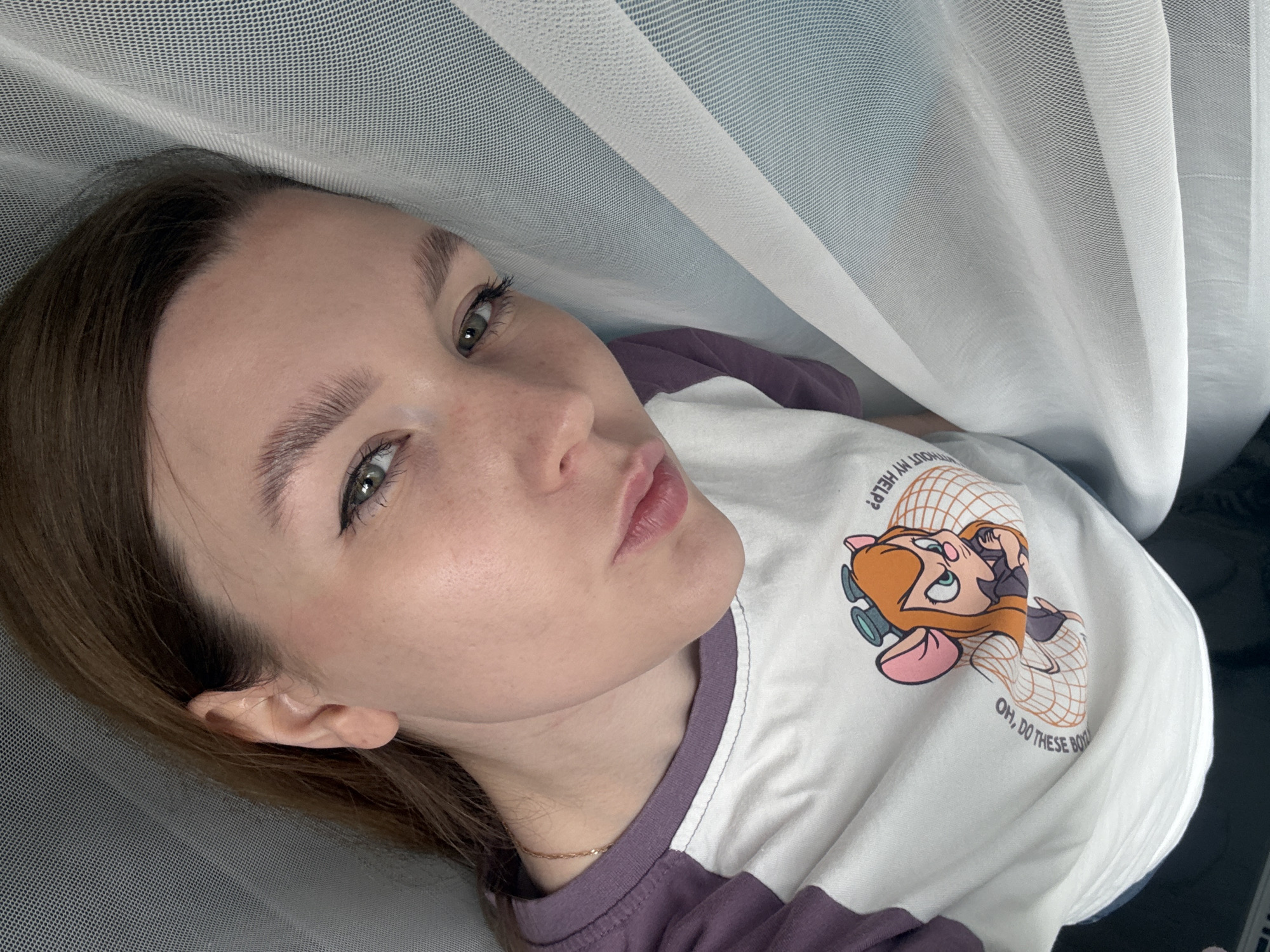 A portrait-style photograph, taken in Izhevsk, Russia, features a young woman lying down indoors under soft, natural lighting. She has fair skin, long brown hair, and striking green eyes, looking directly at the camera with a neutral, slightly pouting expression. Her eyebrows are well-defined and groomed. She is dressed in a casual white t-shirt with purple raglan sleeves, which has a cartoon graphic of a female mouse character, resembling Gadget Hackwrench, wearing goggles. A thin, gold-colored chain necklace is visible around her neck.
The setting is intimate and softly lit, with a sheer, white, net-like fabric or curtain draped over and around her, creating a diffused and ethereal background. The fabric's mesh texture is prominent in the foreground and background.
Visible text on her t-shirt, accompanying the cartoon character, includes partially cut-off phrases that read "...OUT MY HELP?" and "OH, DO THESE BO...". The overall image conveys a relaxed, close-up shot, focusing on her features within a soft, veiled environment.