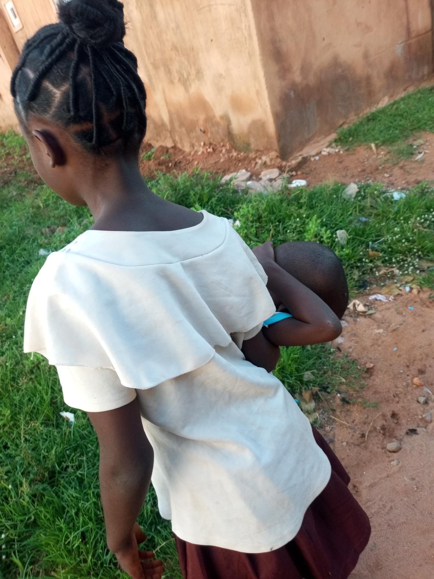 The image shows a young person from behind, with their hair in braids tied into a bun. They are wearing a white top with ruffled sleeves and a dark skirt. The person is holding a smaller child, who is partially obscured. They are standing outdoors on a dirt path next to a grassy area. In the background, there is a building with a rough, plastered wall. The scene appears to be in a rural or semi-urban area. It is not possible to determine the specific location within the city of Tempure, Nigeria, from the provided image.