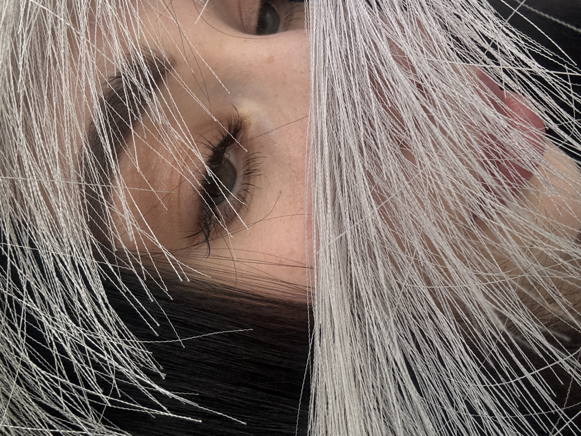 The image presents an artistic, close-up portrait of a person's face, heavily framed and partially obscured by a multitude of fine, white, string-like material. The subject's head is tilted, with their gaze directed upwards and slightly to the left. One eye, with a dark iris and long, dark eyelashes, is clearly visible, while the other eye is partially shown towards the top left. The person has fair skin, dotted with a few freckles.
The white strands are the dominant feature, covering a significant portion of the frame. They are thin, numerous, and fall in various directions, creating a dense, layered texture that partially conceals and partially highlights the facial features. These strands appear to be made of a textile material, resembling very fine yarn or synthetic fibers. On the right side of the image, a subtle hint of the person's pink lips can be seen beneath some of the white strands. Dark natural hair is also visible, providing a strong contrast against the light color of the strands and the skin. The background is generally dark, emphasizing the person and the white material. The image captures a moment of static posing, likely for a photograph, with no specific activity taking place. There are no visible texts or explicit location cues within the image itself. The overall impression is one of intricate detail and textural play.