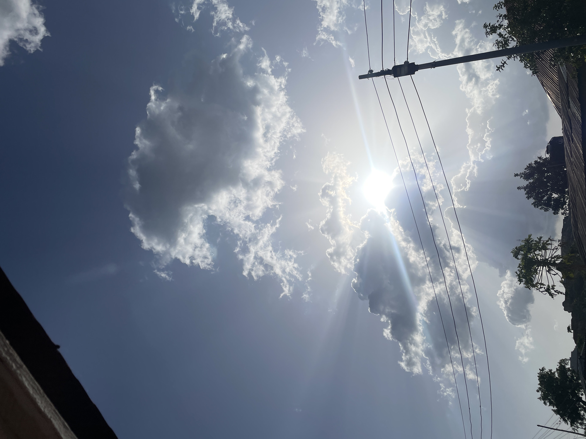 The image is a daytime shot looking upwards at a partly cloudy sky with the sun peeking through the clouds. The sky is a pale blue. The sun's rays are visible. In the upper-right portion of the image, there's a utility pole with several power lines running across the frame. Adjacent to the pole, there is a tall building with some trees and vegetation visible along the edge. The scene is outdoors, possibly in Jos, Nigeria, due to the likelihood of the photo being taken there and the urban elements visible, such as the power lines and buildings.
