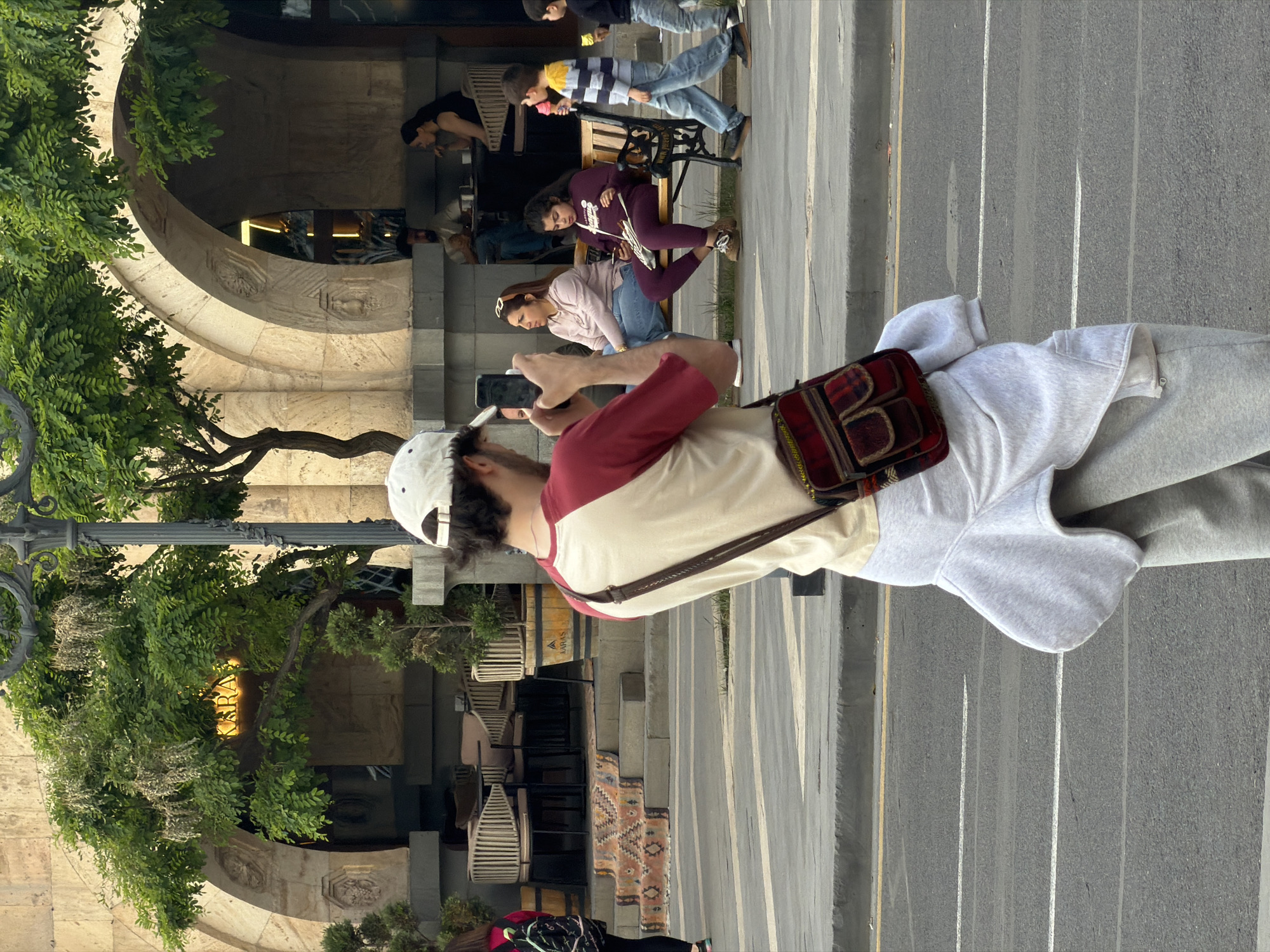 A person is standing on a street in Yerevan, Armenia, holding a mobile phone up to take a picture. The person is wearing a white baseball cap, a red and beige shirt, gray sweatpants, and a crossbody bag with a patterned design. The street is gray with white and yellow lines. Behind the person is a building with arched entrances and a sign that reads "KARAS" partially obscured by foliage. Several people are seated on benches in front of the building. There are tables and chairs visible under the arched entrances.
