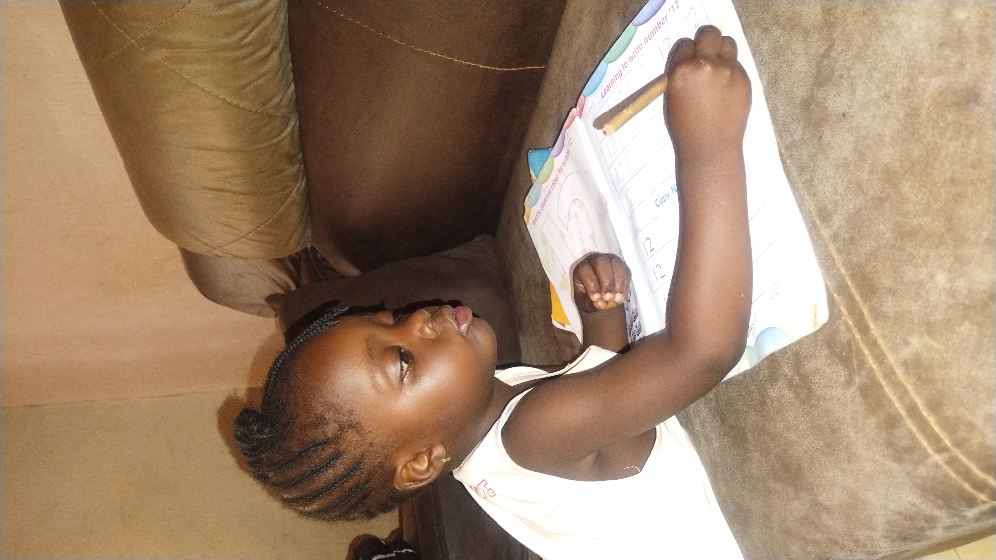 A young, dark-skinned child is seen writing the number "12" in a workbook. The workbook says "Learning to write number '12.'" The child is holding a pencil in their right hand, which is raised above the workbook. The scene appears to be set inside a home with a brown sofa. Based on the presence of English text in the workbook, the scene may be located in Jos, Nigeria, though this cannot be definitively confirmed from the image alone.