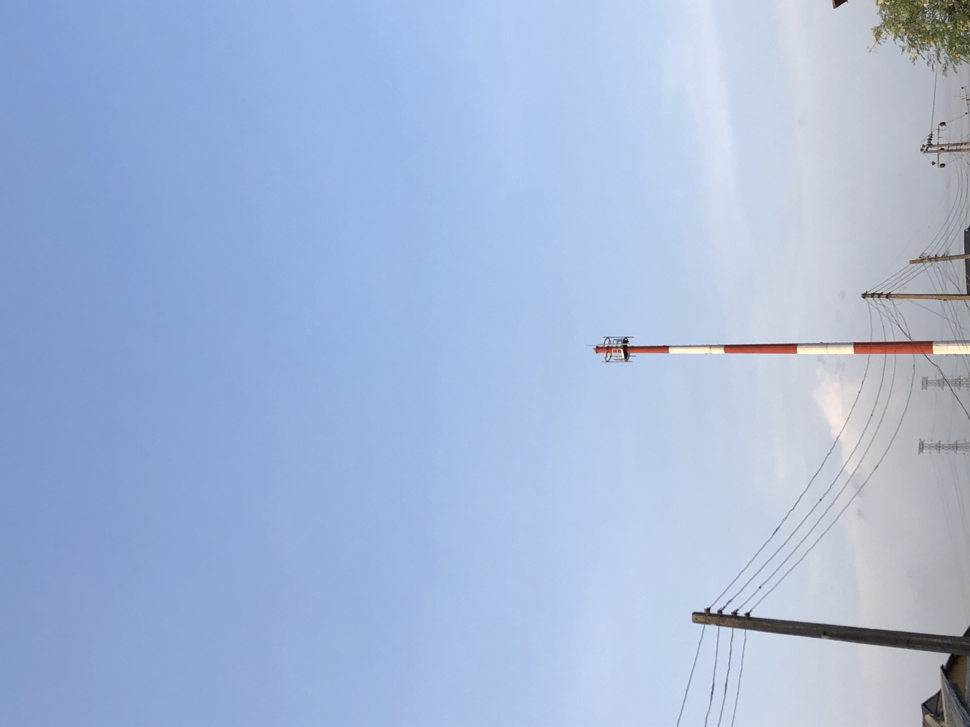 The image shows a clear blue sky with a horizontal red and white striped tower extending from the right side of the frame towards the center. Several electrical poles with wires stretch across the right side of the image. The tower appears to be a communication tower, and the electrical poles suggest an urban or suburban environment. The location is Unknown, Unknown.