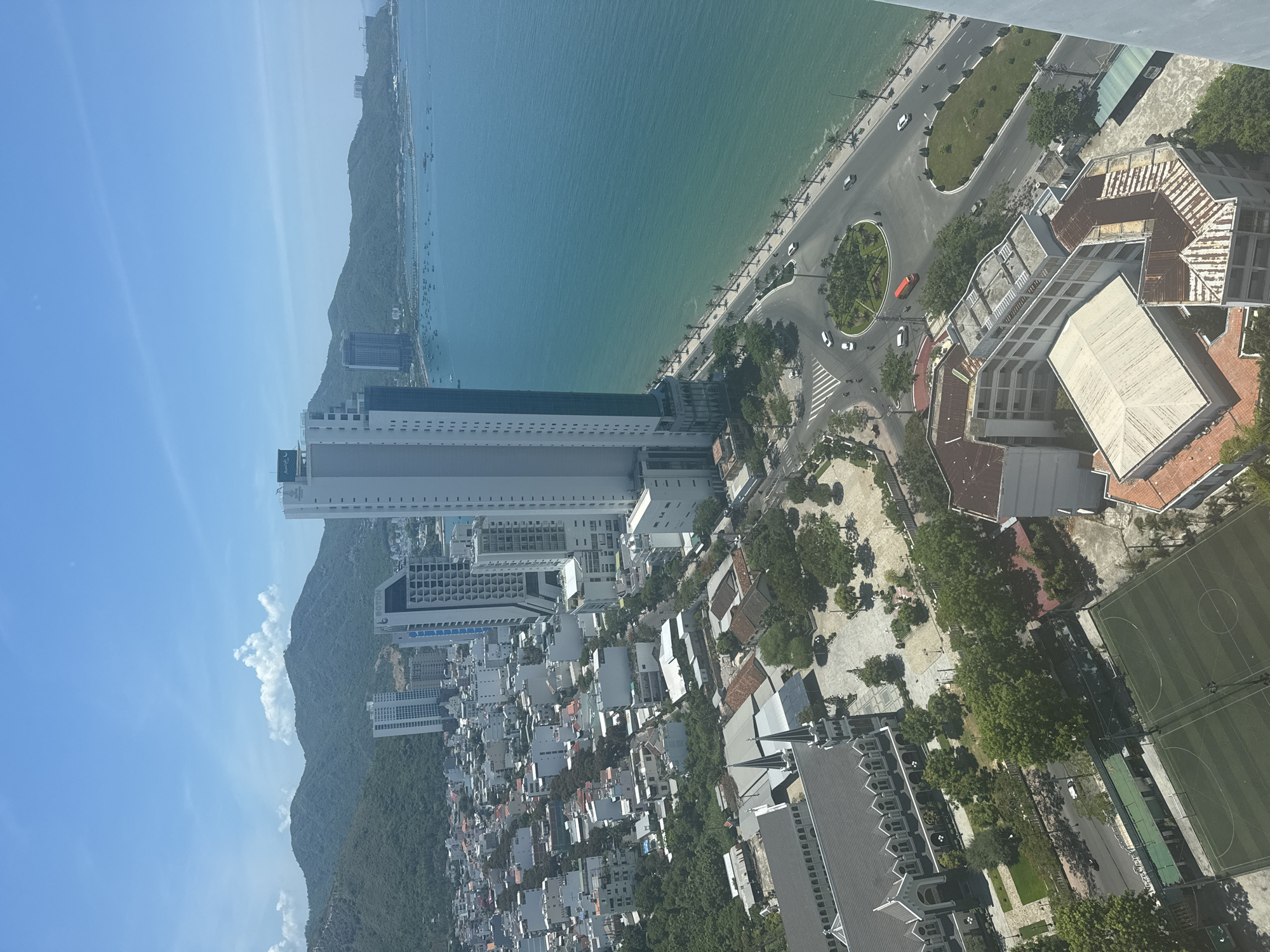 This aerial photograph captures a vibrant coastal city scene, likely in Nha Trang, Vietnam, on a clear, sunny day. Dominating the foreground is a sprawling urban landscape with a mix of architectural styles. A prominent church, identifiable by its gothic-inspired design and twin spires, is situated in the lower left quadrant. Adjacent to it is a green, fenced sports field, possibly a soccer or futsal pitch.
Further into the city, a dense collection of buildings, predominantly residential or commercial, stretches inland. These structures range from low-rise buildings with red-tiled roofs to mid-rise structures. Several modern, tall white skyscrapers punctuate the skyline, with one particularly striking building featuring a blue-tinted glass facade that stands out against the azure sky.
The city gracefully meets the ocean along a curved coastline. A well-maintained road, lined with palm trees, follows the shoreline, with a round-about featuring a central green space. Several vehicles, including cars and motorcycles, are visible navigating the road. The sea itself is a striking turquoise, dotted with numerous small boats, suggesting active fishing or recreational activity. In the distance, lush green mountains form a dramatic backdrop to the city and the sea, with a hazy sky suggesting a warm climate. The overall impression is one of a bustling, modern city integrated with its natural coastal environment.
