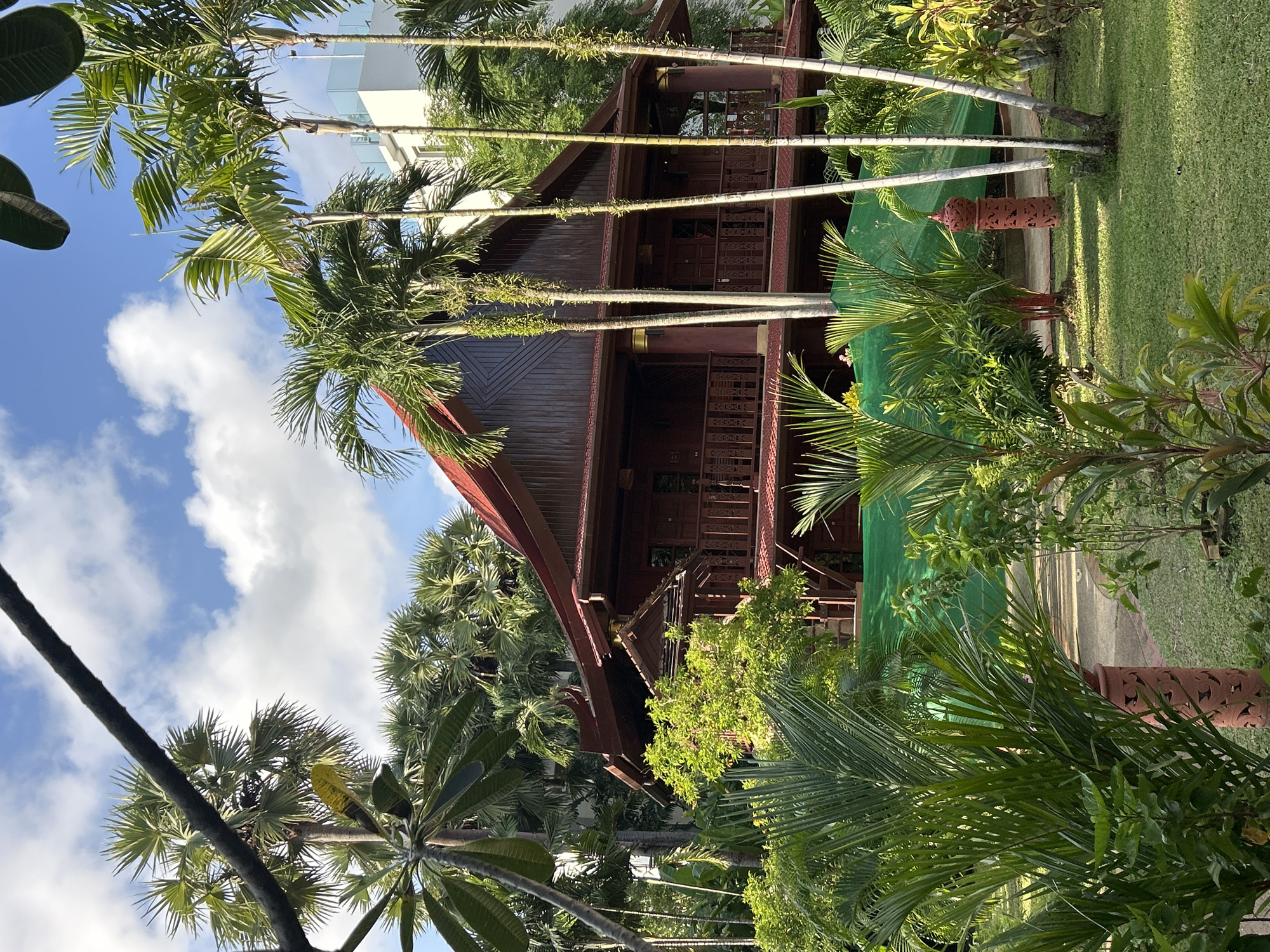 The image depicts a traditional wooden building with a red tiled roof, surrounded by lush tropical vegetation and palm trees. The building appears to be a house or a resort, with balconies and intricate wooden carvings. In the background, a modern white building is visible, suggesting a contrast between traditional and contemporary architecture. The sky is blue with scattered white clouds, indicating a clear day. The foreground is dominated by various types of plants and trees, creating a dense and verdant atmosphere. A green mesh screen is draped over some of the plants, possibly for protection or cultivation. The ground is covered with green grass and a paved pathway. Two decorative terracotta pillars with carved patterns are placed on the lawn. The overall scene is serene and picturesque, with a hint of the bustling city of Patong, Thailand, implied by the presence of modern structures.