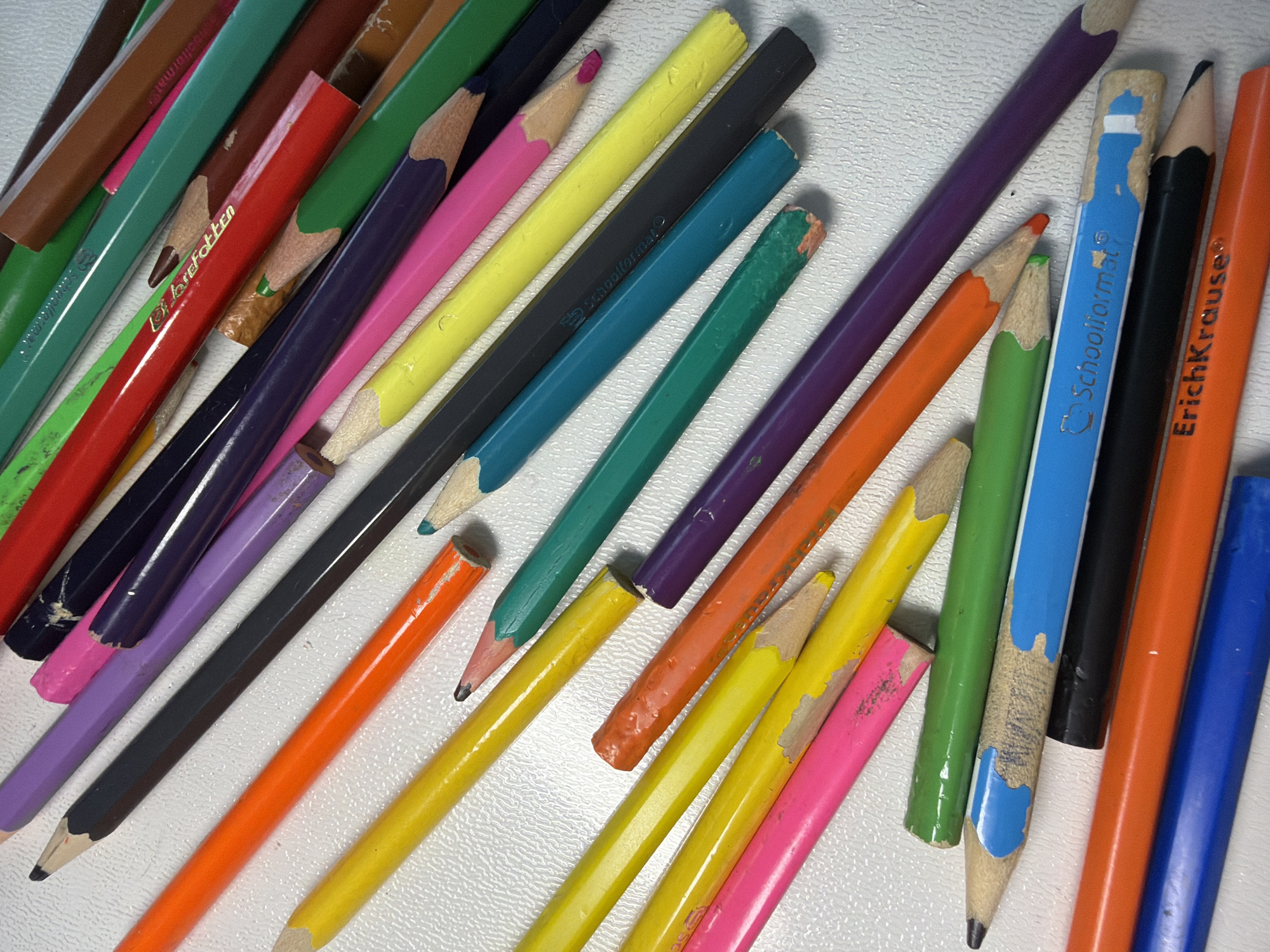 The image shows a collection of colored pencils scattered on a white, textured surface. The pencils are various colors, including brown, green, red, purple, black, yellow, pink, orange, teal, and blue. They appear to be used, with some having worn tips and damaged paint. There is writing or branding visible on several of the pencils, including "Schoolformat" and "Erich Krause." The environment suggests an art studio or crafting space. There is no indication of the location being Votkinsk, Russia, or any people, actions, or interactions in the image.