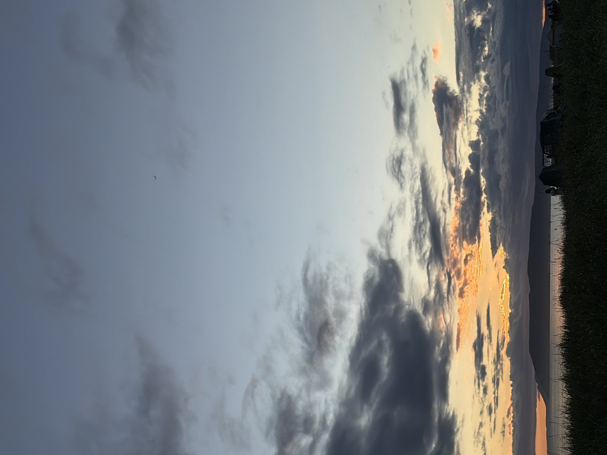 A vertical, wide shot captures a dusky sky with scattered clouds over a body of water. The sky transitions from a pale blue at the top to hues of orange and yellow near the horizon, indicating a sunset. The water is dark and appears calm, with a silhouetted mountainous coastline in the distance. In the foreground, tall grass grows on land, and several tents and figures of people are visible, suggesting a campsite or gathering. A single bird is in flight in the upper portion of the sky. The scene is set in Ozerskoe, Russia.