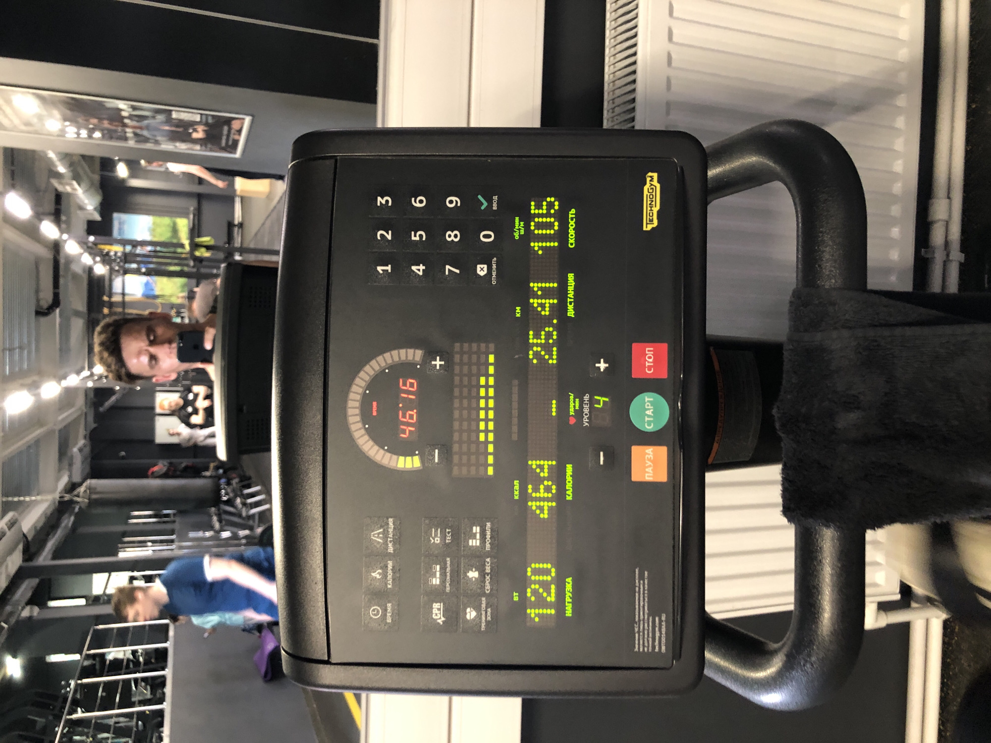 A man takes a selfie in a gym mirror while on an exercise bike. The man is wearing a blue t-shirt and has short brown hair. He is holding a black smartphone up to take the photo. The exercise bike has a digital display showing various statistics, including time, distance, calories burned, and heart rate. In the background, other gym equipment and people can be seen. The gym appears to be modern and well-equipped. The lighting is bright and overhead. The overall impression is that of an active and health-conscious environment. The gym is located in Saint Petersburg, Russia.