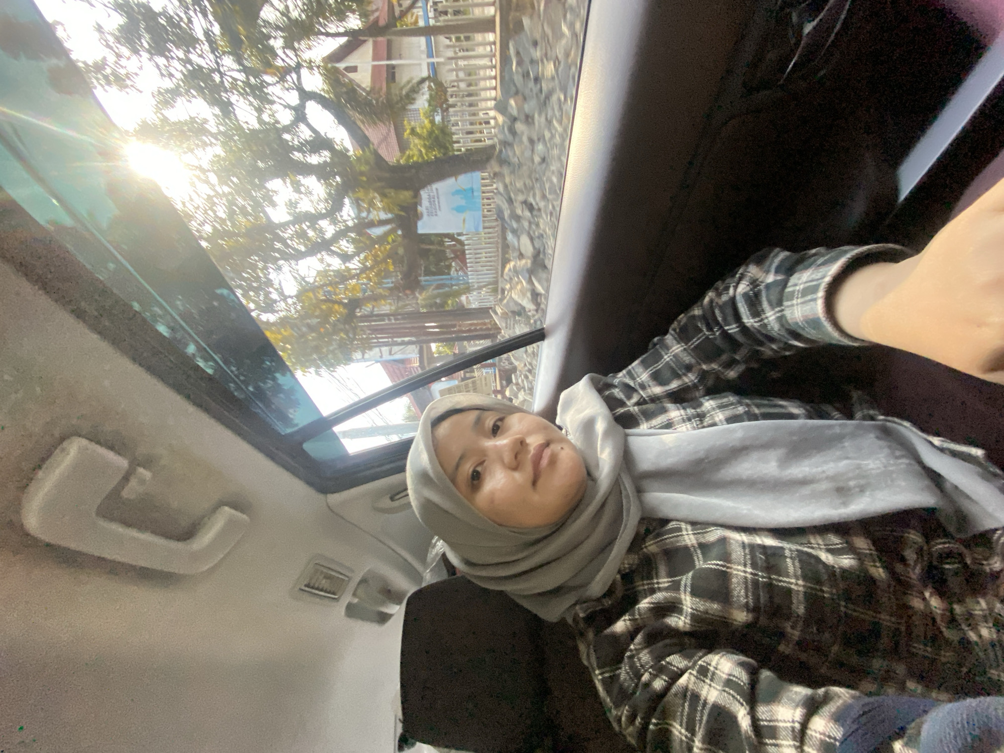 In a selfie taken from inside a vehicle, a young woman wearing a gray hijab and a black and white plaid shirt looks towards the camera. The interior of the car is visible, with a handle on the ceiling and a vent. Sunlight streams through the window, illuminating trees and a building outside. The scene suggests a casual, everyday moment captured during a car ride. While the image doesn't explicitly state the location, the user has specified the city of Banjarmasin, Indonesia.