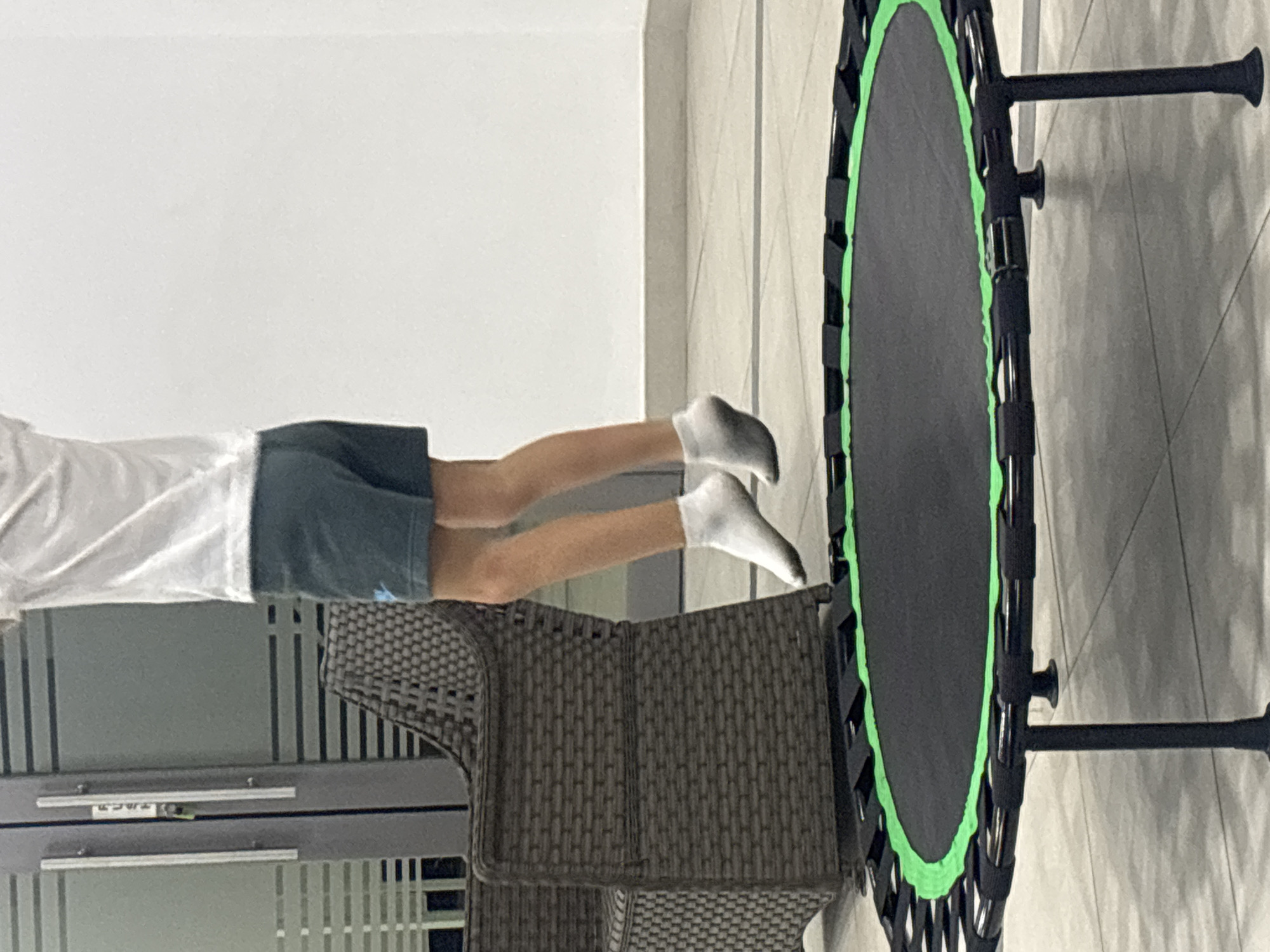 A person is captured mid-air, jumping on a trampoline. The person is wearing a white t-shirt, dark shorts, and white socks. The trampoline is black with a green edge and has black legs. In the background, a woven chair is visible next to a door with frosted glass panels. The scene appears to be an outdoor or semi-outdoor space, with a tiled floor and a white wall. The environment suggests a modern setting, possibly in a residential area or a recreational facility. The image does not provide enough context to definitively place it within Dubai, United Arab Emirates, or any specific city.