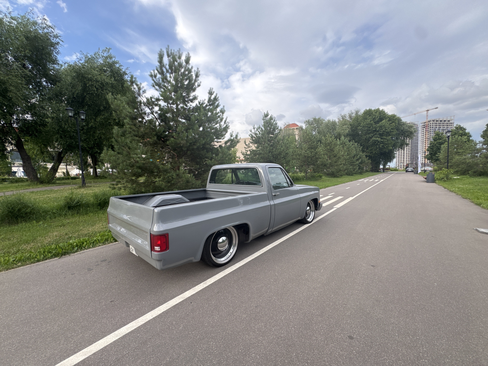 A grey pickup truck is parked on an asphalt path in a park on a cloudy day. The truck is in good condition and appears to be a classic model. The path is lined with green grass and trees. In the background, there are several tall buildings under construction, one with a crane. The sky is overcast with some clouds. The scene suggests a relaxed, possibly recreational atmosphere in an urban park setting. No people are visible in the image. The time of day appears to be daytime.