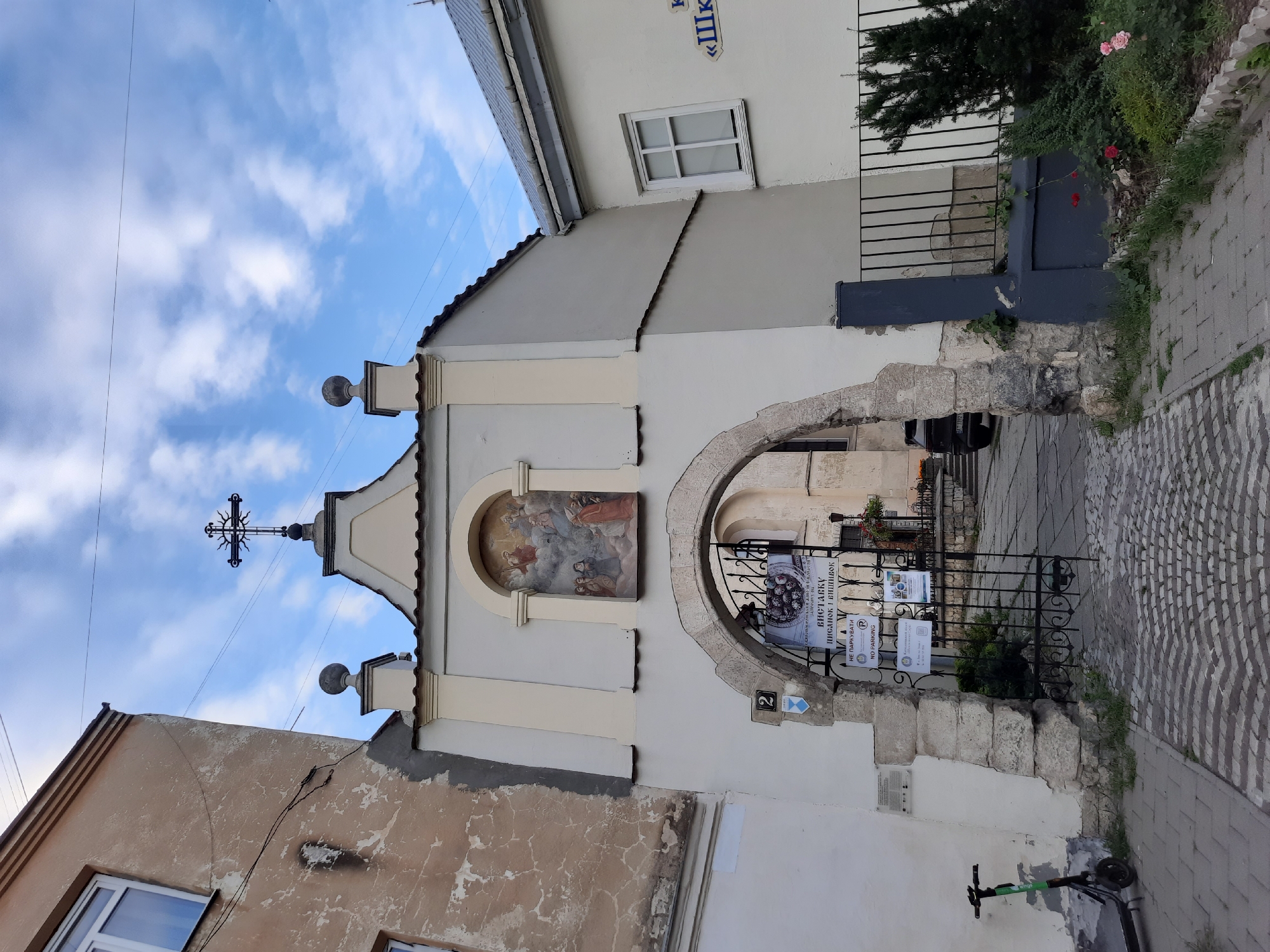 The image captures a view of an arched stone entrance leading into a courtyard with a building adorned with a religious fresco and a cross. To the left, a weathered, multi-story building with visible peeling plaster stands adjacent to the arched entrance. The entrance itself is framed by a large, rough-hewn stone archway, partially covered by a decorative metal gate.
Above the arch, a fresco depicts a religious scene with figures in the sky. A cross with a sunburst design is mounted on the peak of the building. On the gate, several signs are posted, one featuring what appears to be an exhibit announcement in Ukrainian, and another with a "No Parking" symbol and text.
To the left of the entrance, a gray electric scooter is parked on a cobblestone sidewalk. The cobblestone path extends into the courtyard, which is visible through the gate. Beyond the arch, a dark-colored car is parked in the courtyard, and steps lead up to another building.
On the right side of the image, a white building with a window and a dark gray, fence-like structure is visible. A small garden with roses and evergreen bushes is situated in front of this fence. The sky is blue with scattered white clouds. The overall scene suggests a historical urban setting, likely in Lviv, Ukraine, given the architectural style and the Ukrainian text on the signs.