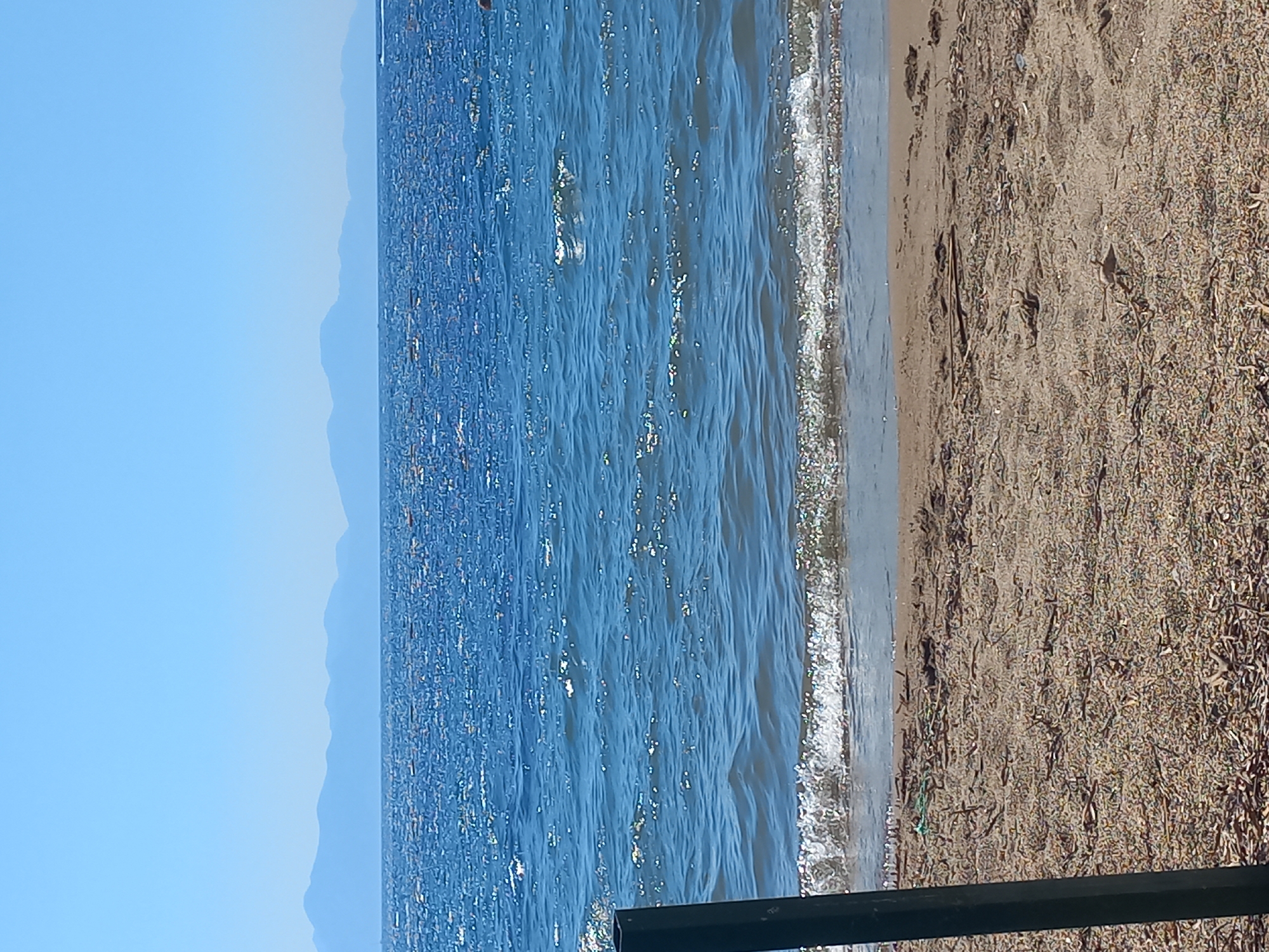 The image depicts a tranquil beach scene under a clear blue sky. In the foreground, a sandy beach is scattered with small pebbles and dried vegetation, with a dark metal post on the left side. Gentle waves lap at the shore, creating a foamy white line where they meet the sand. The sea stretches out to the horizon, its surface shimmering with sunlight, and in the distance, a hazy mountain range is visible. The overall impression is one of peacefulness and natural beauty. While the specific location in Bodrum, Turkey, is not visually identifiable, the landscape is consistent with coastal scenery found in such areas.