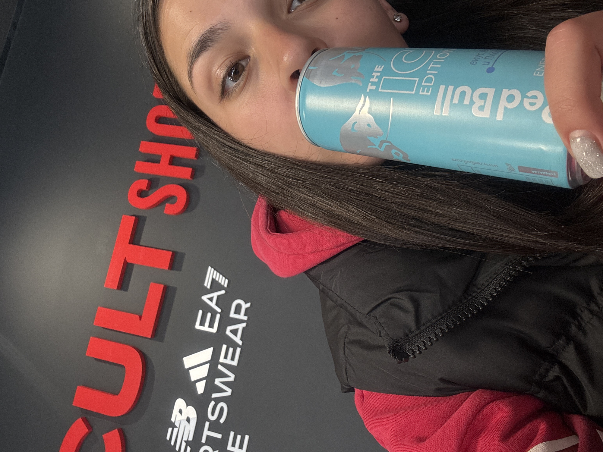 A woman holds a blue Red Bull energy drink can labeled "THE IC EDITION" near her face, with the "CULT SHOP" sign and logos of Adidas, EA7, and New Balance visible in the background. The location is Gabrovo, Bulgaria.