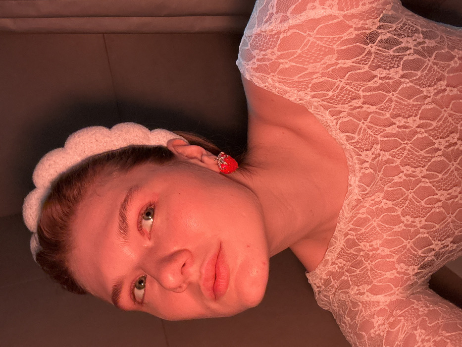 An angled, close-up indoor portrait features a young adult female under reddish illumination. She wears a white, soft, scalloped headband and a white, long-sleeved lace top with a patterned open weave. A prominent red, strawberry-shaped earring, detailed with green and silver accents, is visible on her right earlobe. Her eyes are open, and her gaze is directed upwards and to the right. The background is dark and indistinct, exhibiting subtle color variations from the reddish light. No specific location within Kazan, Russia, is discernible from the visual information.