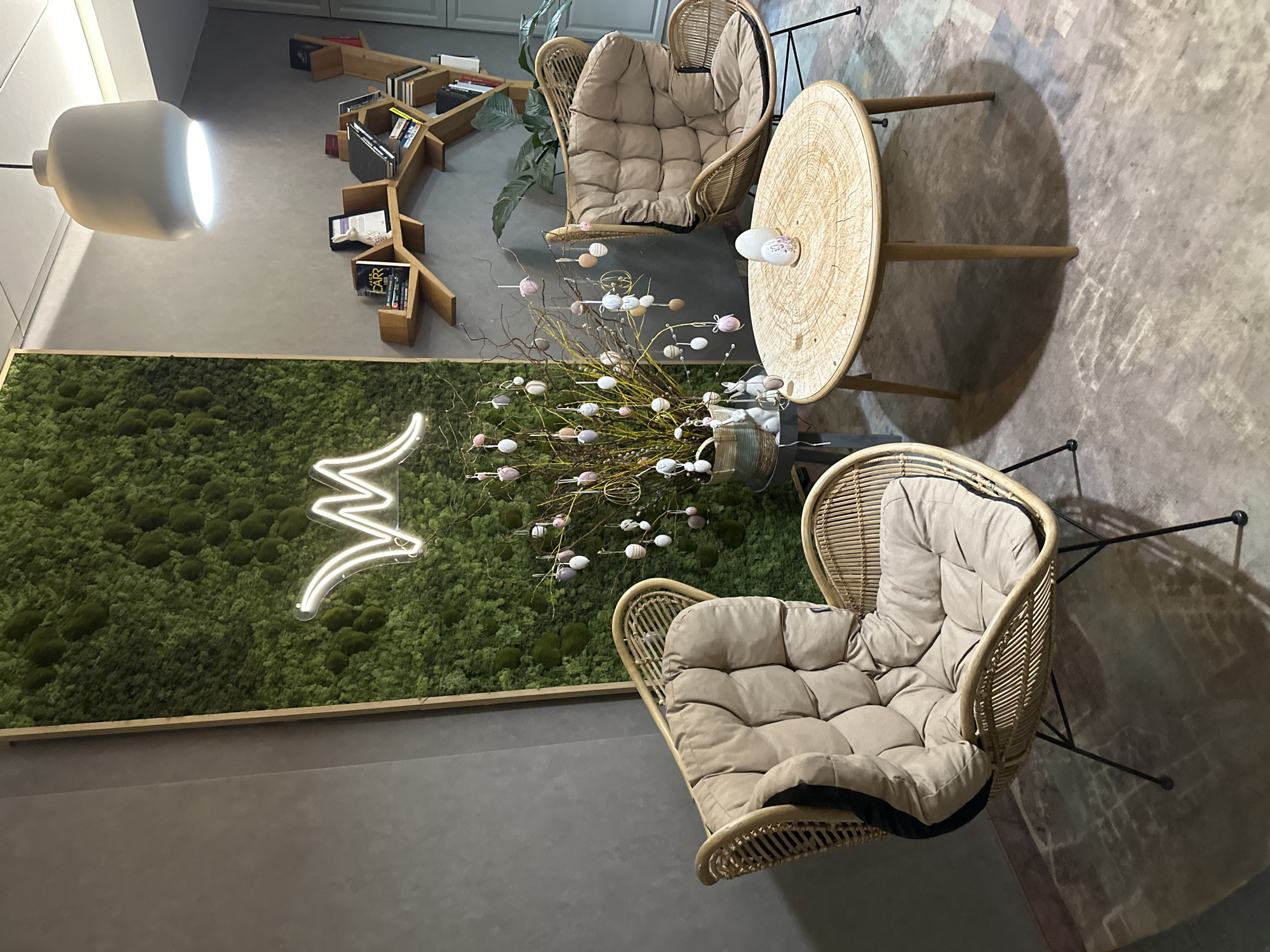 The image depicts a modern, stylized interior, likely a cafe or lounge area. Two wicker chairs with plush cushions are arranged around a round wooden table. On the table is a decorative vase with branches adorned with Easter egg ornaments, suggesting a seasonal theme. A white ceramic object rests on the table as well.
Behind the seating arrangement, a vertical garden wall is visible, decorated with a neon sign in the shape of a stylized letter "M". To the left, a modern lamp hangs from the ceiling, providing illumination. In the background, a bookshelf with a unique, branching design holds books.
The floor appears to be made of concrete or a similar industrial material. The overall aesthetic is a blend of natural elements and contemporary design.
The location is inferred to be Wrocław, Poland based on the initial prompt that requests this detail. However, there are no discernible visual cues within the image to definitively confirm this location.