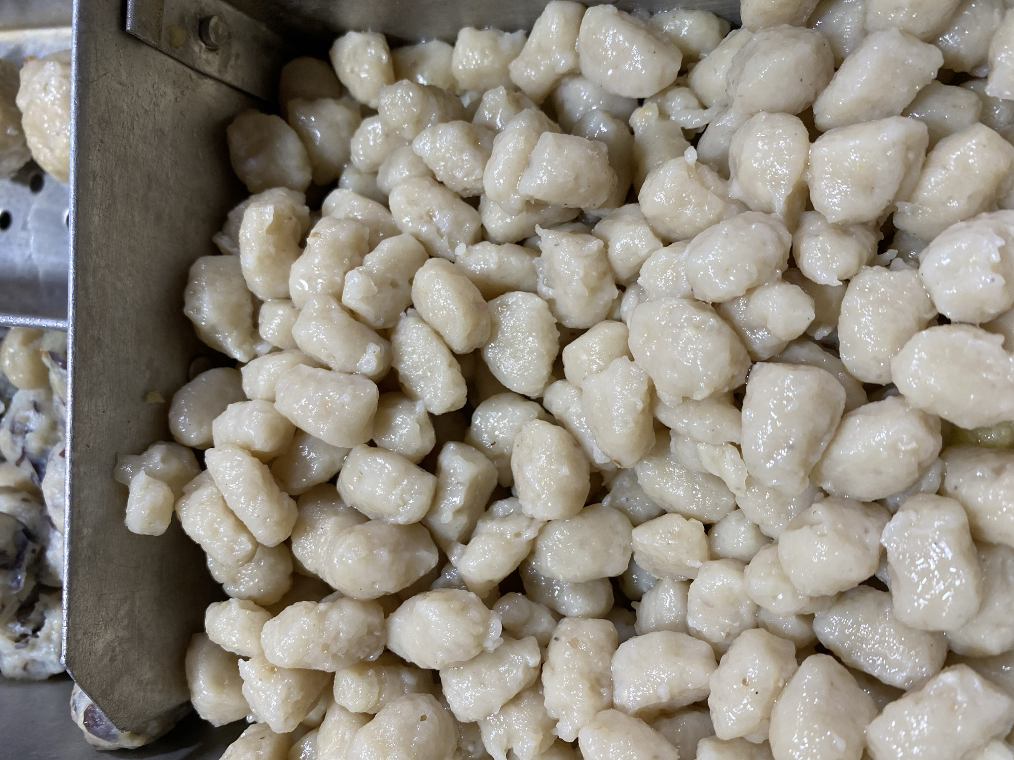 The image shows a close-up, overhead view of a metal container filled with a large quantity of small, irregularly shaped, pale, cooked food items, likely meatballs or dumplings, suspended in a light-colored liquid or sauce. The food items are dense and appear to have a smooth texture. The container is made of brushed metal and is positioned at an upward angle, revealing more of the contents towards the bottom of the frame. In the background, another metal container is partially visible, containing a similar type of food. The lighting is bright and casts subtle shadows on the food items, highlighting their rounded shapes and slight sheen. The overall impression is of food being prepared or served in a commercial setting, possibly a market or restaurant kitchen. This scene is likely located in a food stall or eatery within the city of Kasihan, Indonesia, known for its diverse culinary offerings.