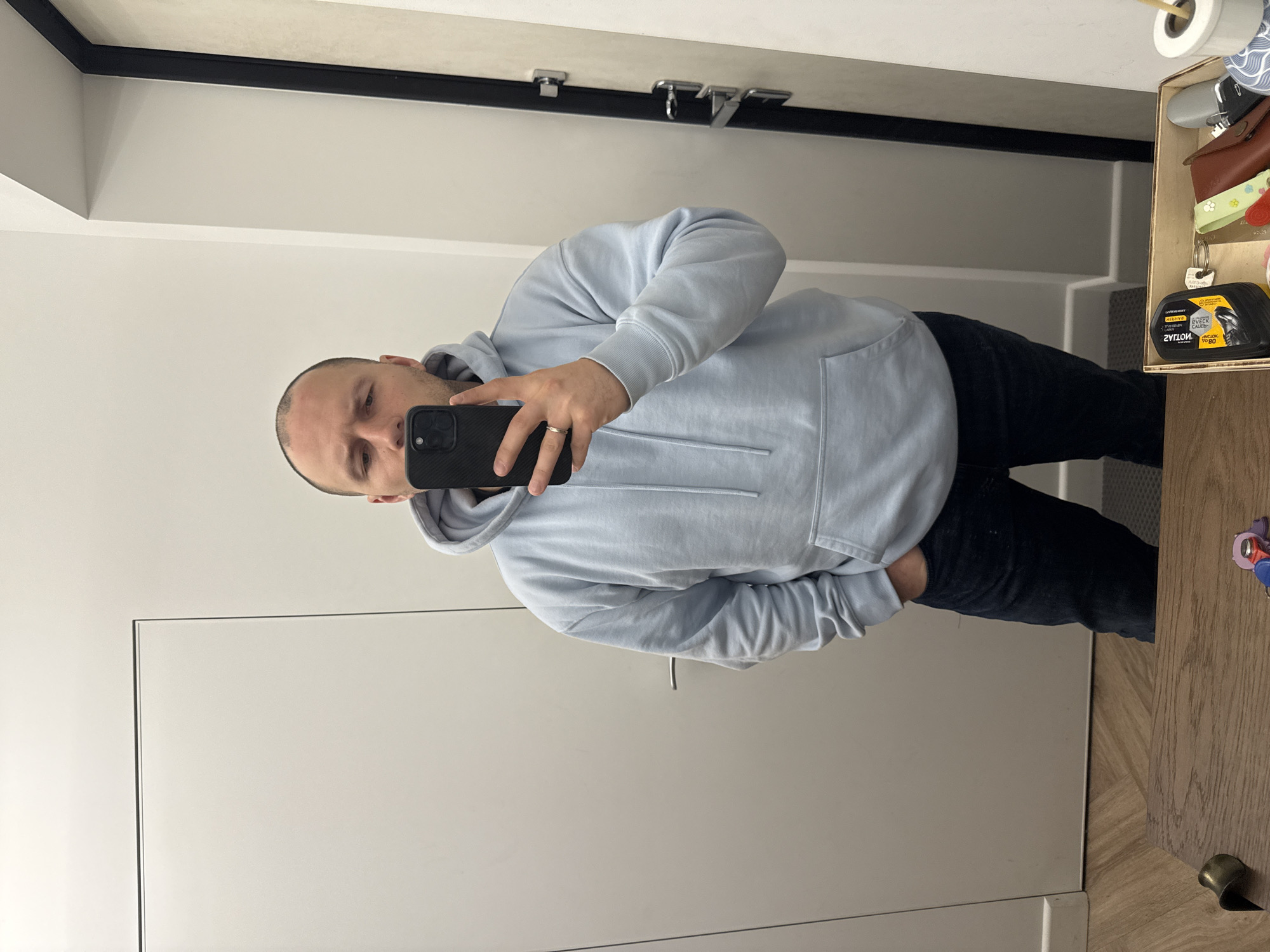 A man takes a selfie in front of a mirror. He wears a light blue hoodie and dark jeans. The background shows a white door and part of a wooden table with keys and other items. The location cannot be determined from the image.