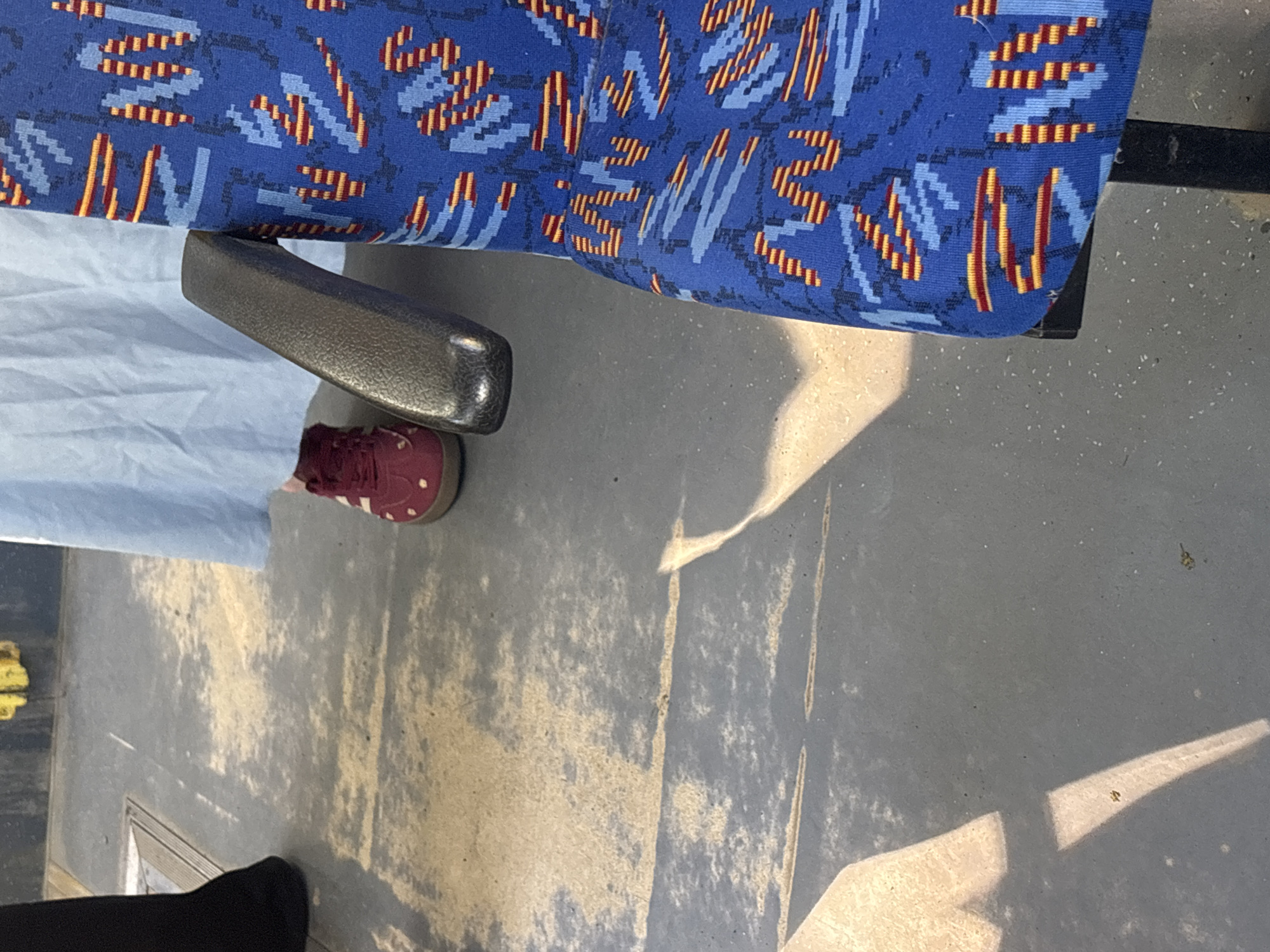 The image shows an interior view, possibly of a bus or other public transportation seating arrangement. A blue seat with a patterned design of blue, orange, and yellow shapes dominates the top of the frame. A gray armrest is adjacent to the seat. Part of a person is visible, wearing a light blue garment and a burgundy-colored shoe with white stripes. The floor is a light gray, with shadows cast by the light streaming in. In the bottom left corner, a portion of a black pant leg is visible. The scene appears to be within a public transportation vehicle, but without further context, the exact location within Saint Petersburg, Russia, cannot be determined.