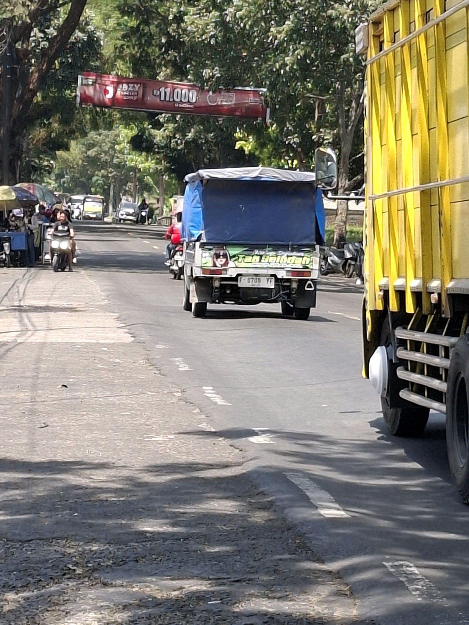 The image captures a street scene in Cikahuripan, Indonesia, on a sunny day with dappled shadows on the asphalt. A large yellow truck dominates the right side of the frame, its side panel showing vertical yellow slats and horizontal cables. In the center of the road, a small white truck with a blue tarp cover is moving away from the camera. It has a license plate reading "F 0709 YF" and a sticker on the back featuring a woman wearing sunglasses. To the left of the truck, several motorcycles and a yellow bus are visible. One motorcycle rider, wearing a black shirt, is in the foreground on the left side of the road. Further back, a person in a red shirt is on another motorcycle. The left side of the road features a sidewalk lined with small stalls under colorful umbrellas. A red banner with white text and the number "11.000" is suspended across the road in the background, partially obscured by trees. The environment appears to be a busy commercial area with moderate traffic.
