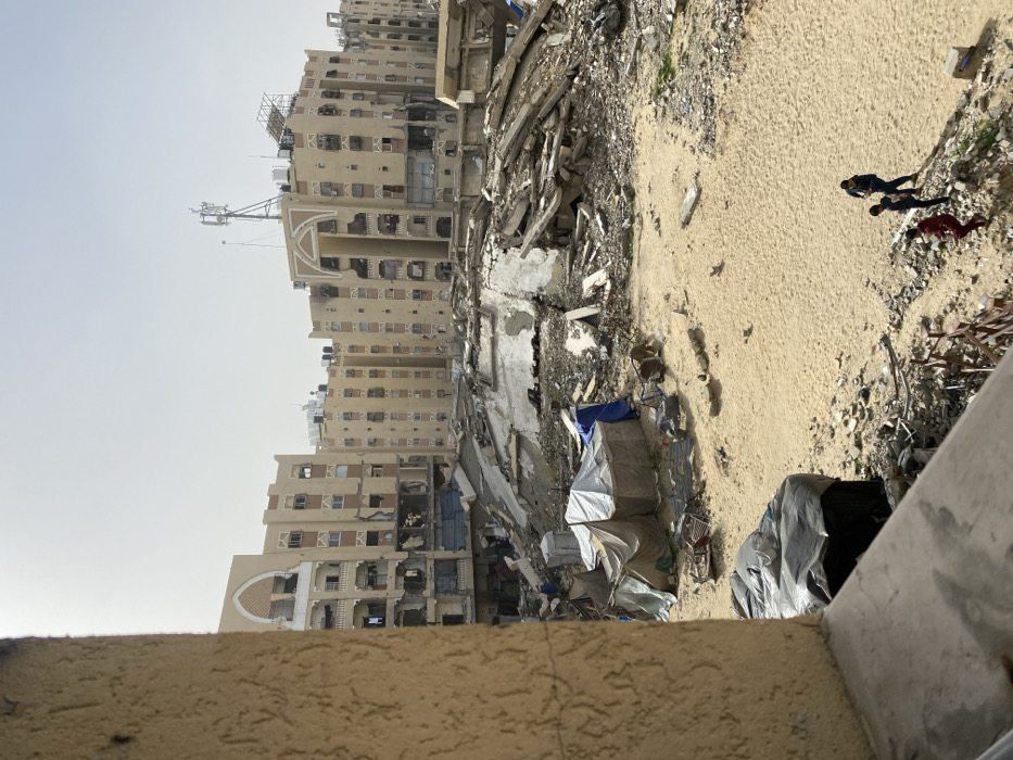 The image displays an extensively damaged urban environment in Khan Yunis, Palestinian Territory, viewed from an elevated position. A light-brown, textured building wall partially frames the left foreground. The midground consists of a large, sandy area covered with scattered debris. On the left midground, several makeshift tents, constructed from light-colored and blue tarpaulins, are erected. A significant pile of concrete rubble and twisted rebar, remnants of destroyed structures, dominates the central midground. Three individuals, appearing to be children, walk across the sandy terrain in the lower right portion of the scene. In the background, multiple multi-story residential buildings, tan in color with arched window frames and entrances, exhibit widespread structural damage, including numerous impact holes, missing wall sections, broken windows, and darkened facades. Rooftops feature black water tanks, communication antennas, and scaffolding. The sky is clear.