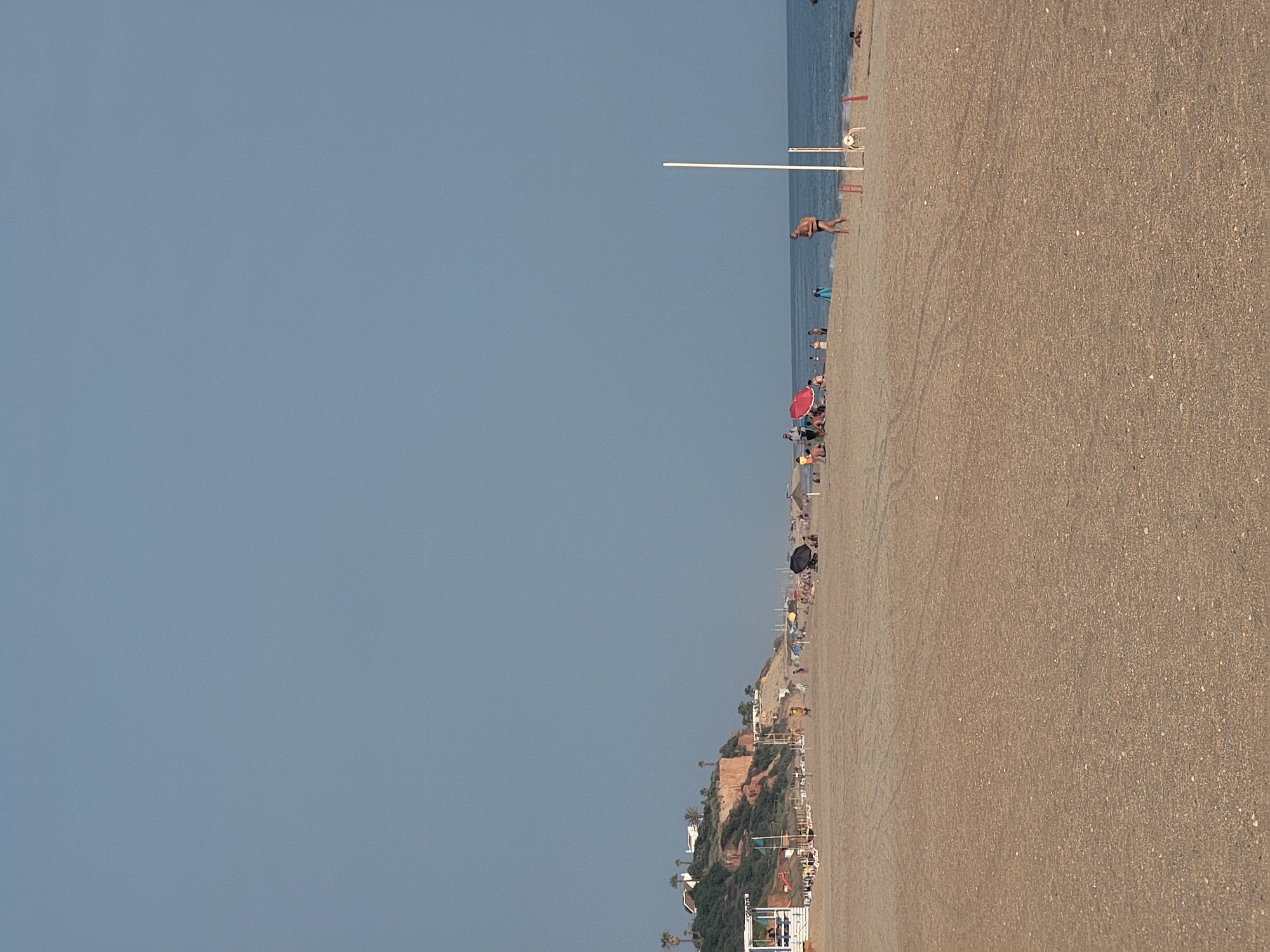 The image depicts a wide, sandy beach in Quarteira, Portugal, under a clear, pale blue sky. The foreground is dominated by the textured sand, marked with tire tracks. In the mid-ground, a gentle slope leads to the water, where a few people are visible. The sea is calm and appears a deep blue. Along the shoreline, scattered beach umbrellas and a white lifeguard tower are present, along with groups of sunbathers and individuals walking. To the left, a rocky cliffside covered with greenery and dotted with white buildings and palm trees overlooks the beach. A thin, white pole stands vertically near the center of the image, possibly a marker or part of a beach facility. A man in dark swim trunks is walking towards the sea in the mid-right portion of the frame, and a dog is visible on the sand to the right of the pole. The scene conveys a typical summer day at a moderately populated beach.