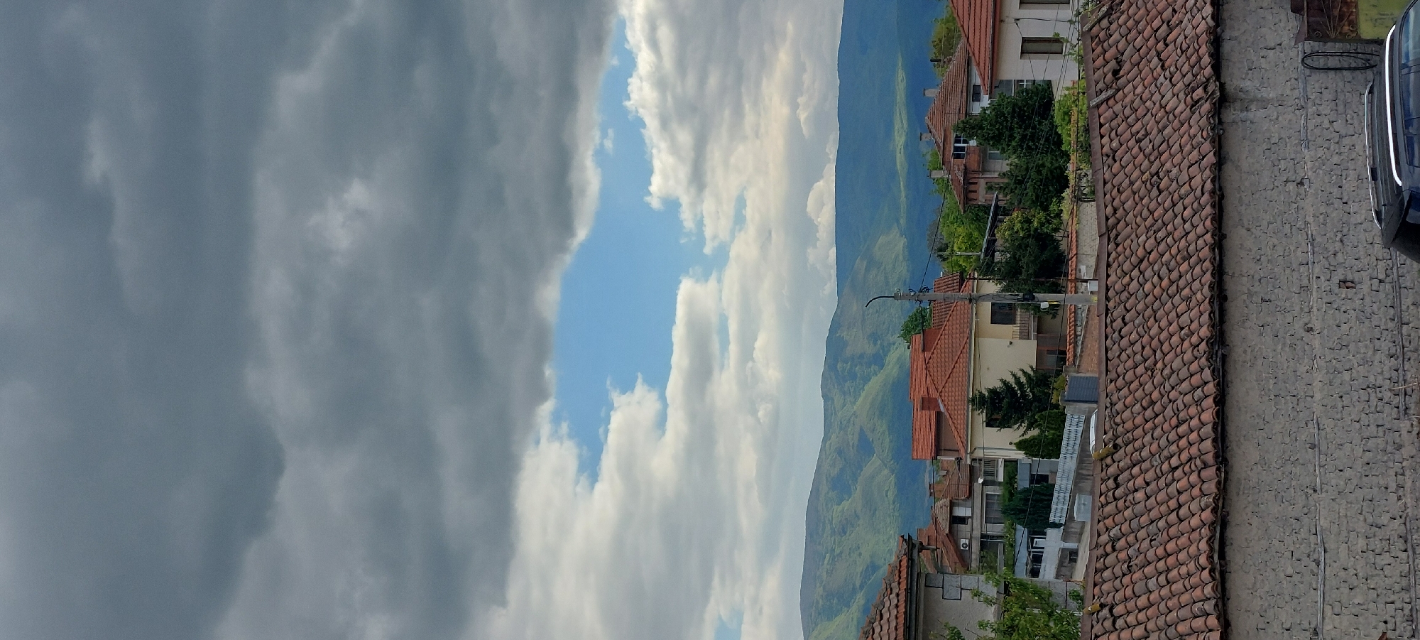 The image depicts a view in Yagodovo, Bulgaria, showing a scene under a partly cloudy sky. The sky is divided into dark, ominous clouds on the left and lighter, partly cloudy skies with patches of blue on the right. Below the sky is a mountainous landscape that is covered in green vegetation. In the foreground, there is a row of buildings with red tile roofs, typical of the area. A cobblestone pathway and a dark-colored car are visible in the lower right corner, indicating a street or roadside view. Electrical poles and wires can be seen extending along the street. No people are visible in the scene. The image appears to have been taken from an elevated position, providing a comprehensive view of the landscape and cityscape.