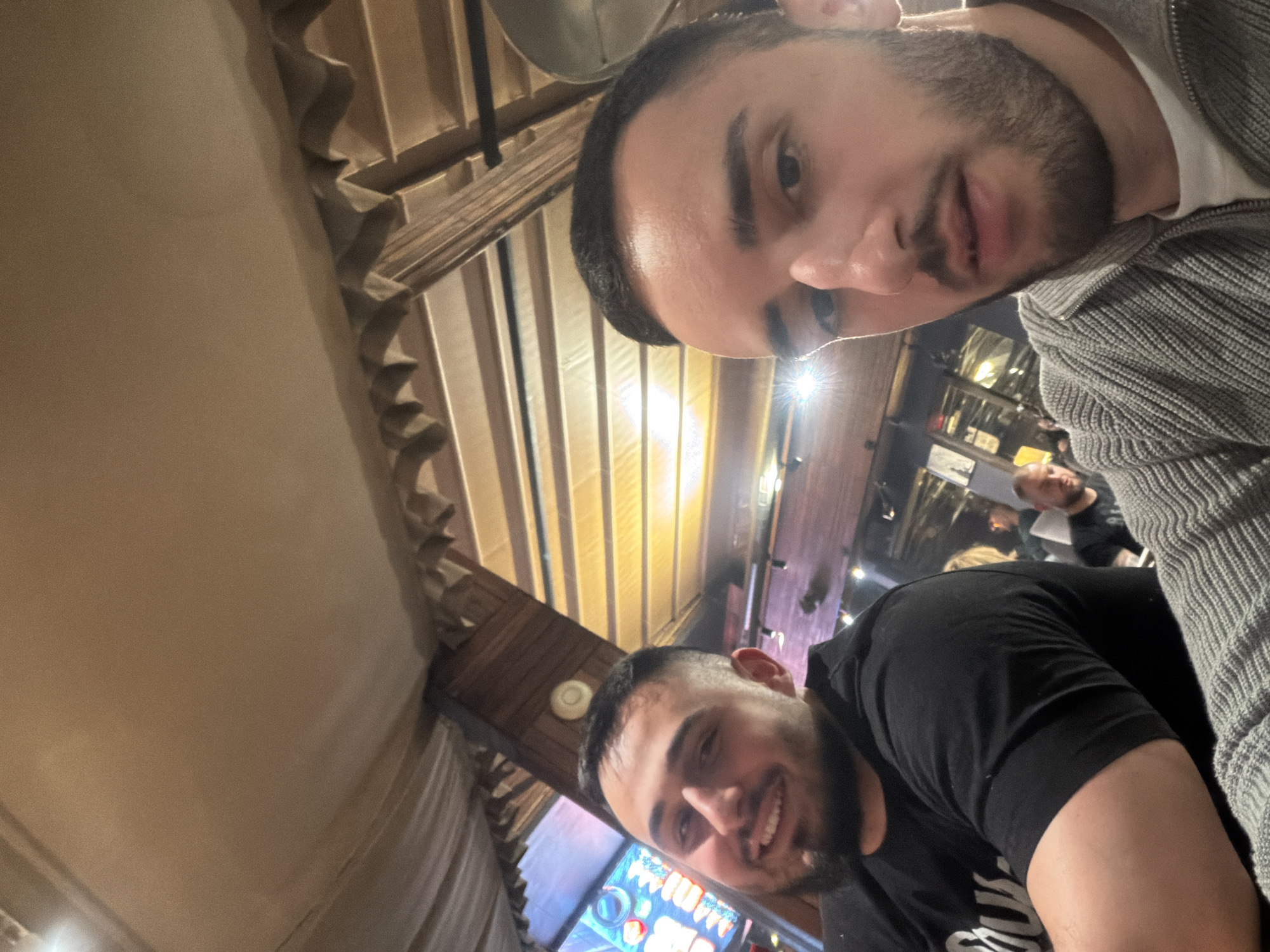 Two men are taking a selfie in a restaurant. The man on the left wears a black T-shirt and smiles. The man on the right wears a grey knit sweater. The restaurant's interior features a wooden ceiling with exposed beams and decorative lighting. Other people are visible in the background. The restaurant is in Plovdiv, Bulgaria, and the photo was likely taken from a table.