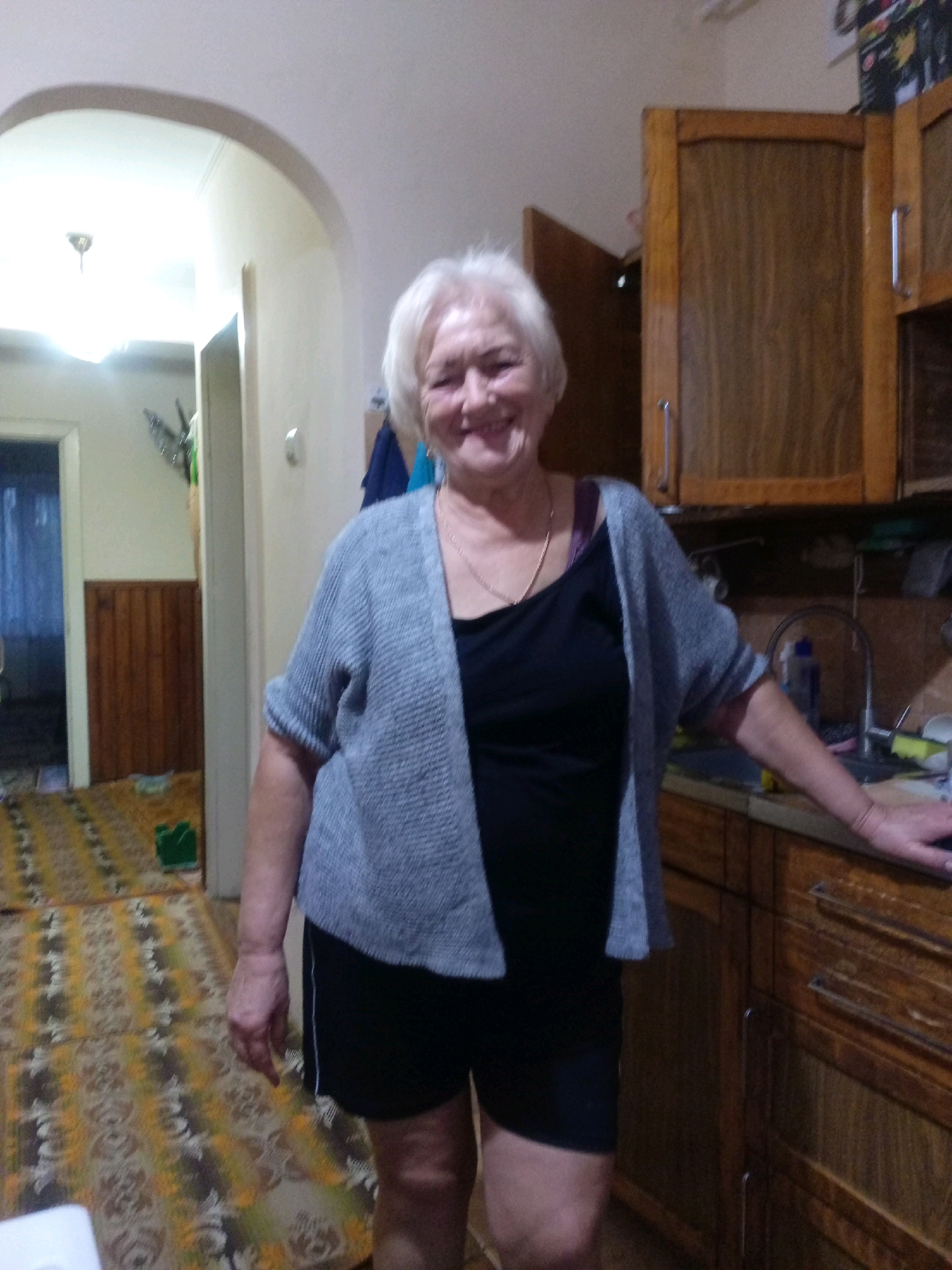 An elderly Caucasian woman with short, blonde hair and fair skin is standing in what appears to be a kitchen or utility area. She is wearing a gray knitted cardigan over a black tank top and black shorts. A thin gold necklace is visible around her neck. She is smiling at the camera. To her right are wooden cabinets with metal handles, and a countertop with a sink and a blue bottle. To her left, an arched doorway leads to a hallway with a patterned rug on the floor. The lighting in the image is dim, suggesting it may be indoors in the evening or in a poorly lit area. The overall scene conveys a casual, domestic setting. The location is identified as Tiachiv, Ukraine.