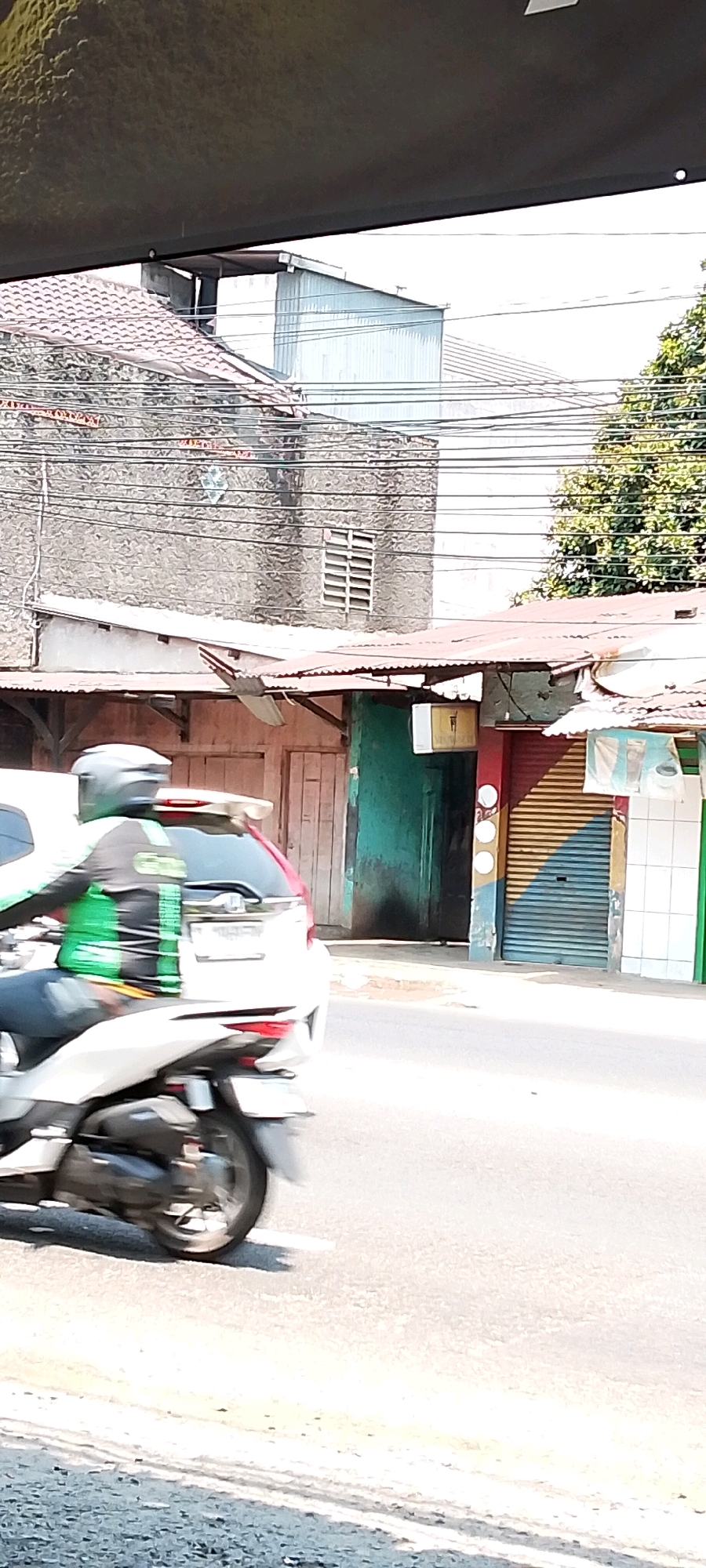 A person wearing a dark grey helmet and a green and black uniform is riding a white motorcycle on a paved road. The motorcycle is moving from left to right. Behind the motorcycle, there is a white car. The background consists of buildings with corrugated metal roofs and stone walls. One building has a large, faded green door. Another building features a rolling metal door with horizontal yellow and blue stripes. Overhead power lines crisscross the scene. The environment suggests a street in Depok, Indonesia, characterized by its urban architecture and infrastructure. The lighting indicates it is daytime, with shadows cast on the road.